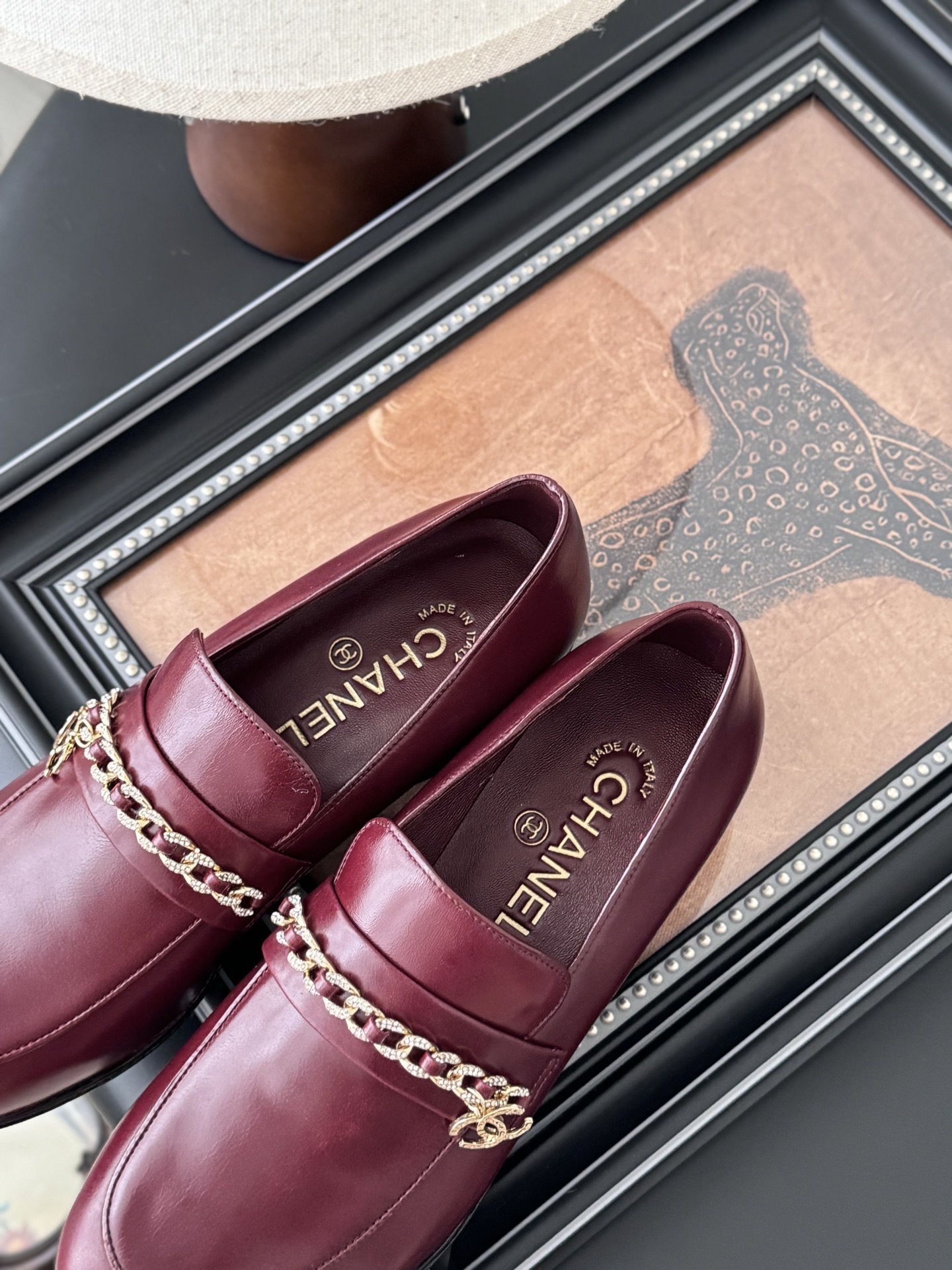 Pair of burgundy Chanel leather loafers with gold chain detail and block heels next to a black Chanel shoe box. - Hình 8