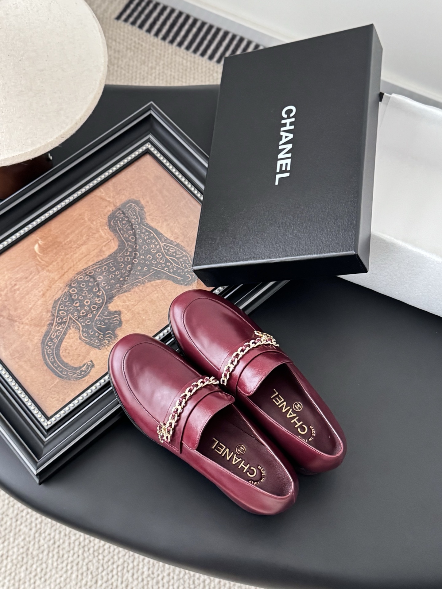 Pair of burgundy Chanel leather loafers with gold chain detail and block heels next to a black Chanel shoe box. - Hình 7
