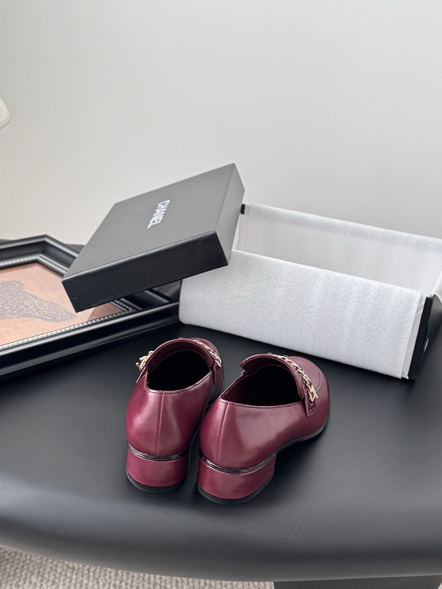 Pair of burgundy Chanel leather loafers with gold chain detail and block heels next to a black Chanel shoe box. - Hình 6