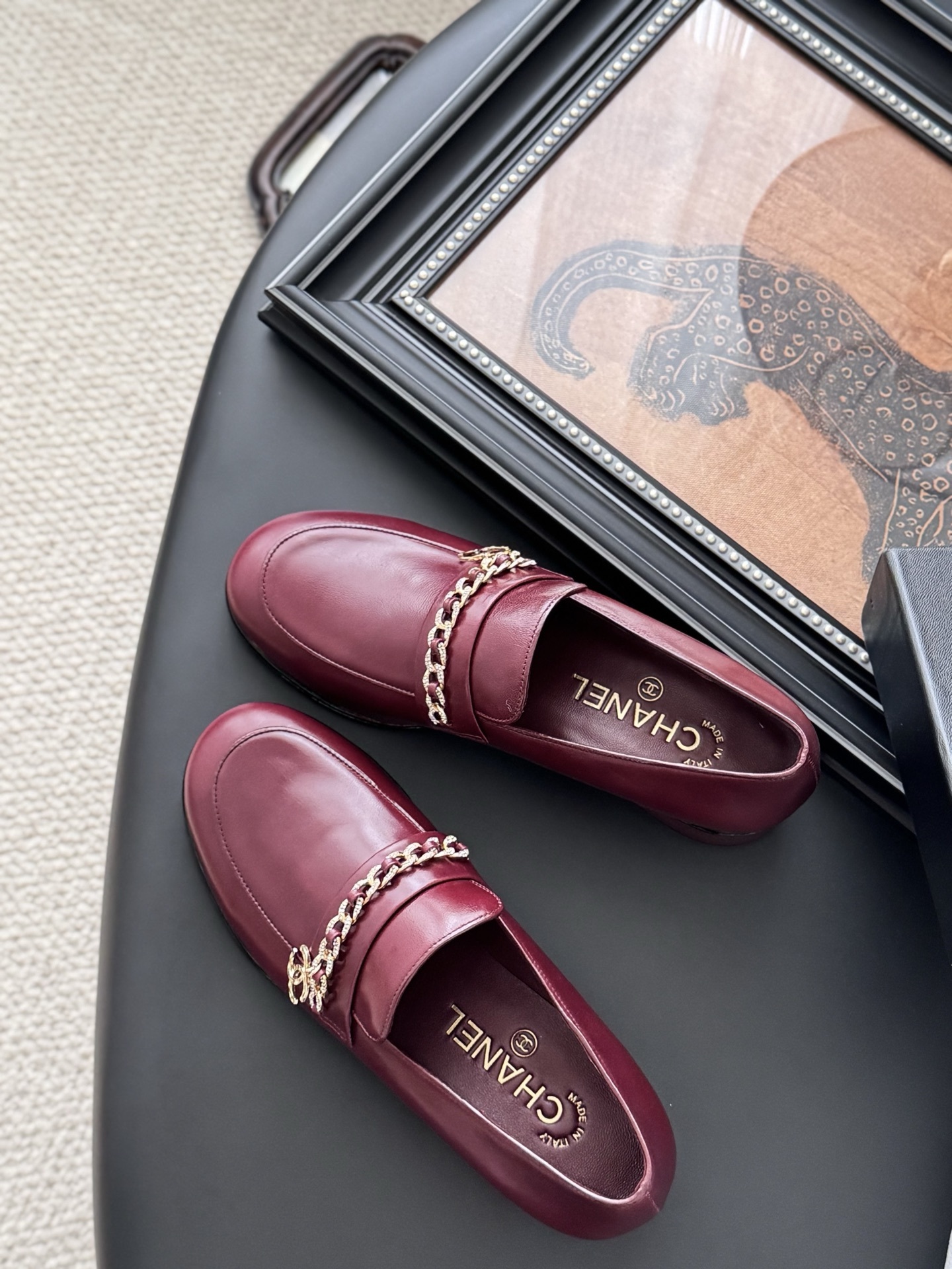 Pair of burgundy Chanel leather loafers with gold chain detail and block heels next to a black Chanel shoe box. - Hình 5