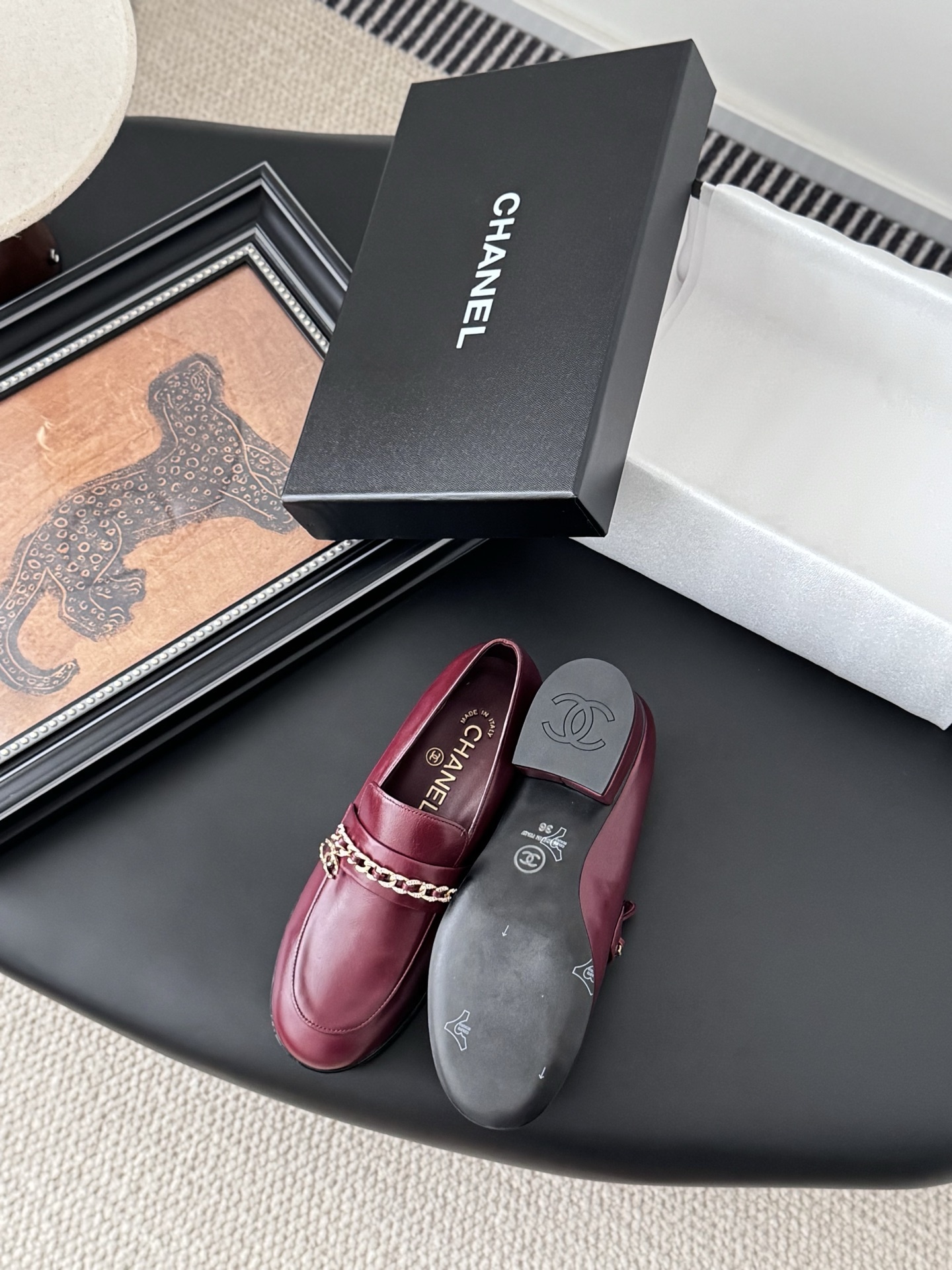 Pair of burgundy Chanel leather loafers with gold chain detail and block heels next to a black Chanel shoe box. - Hình 4
