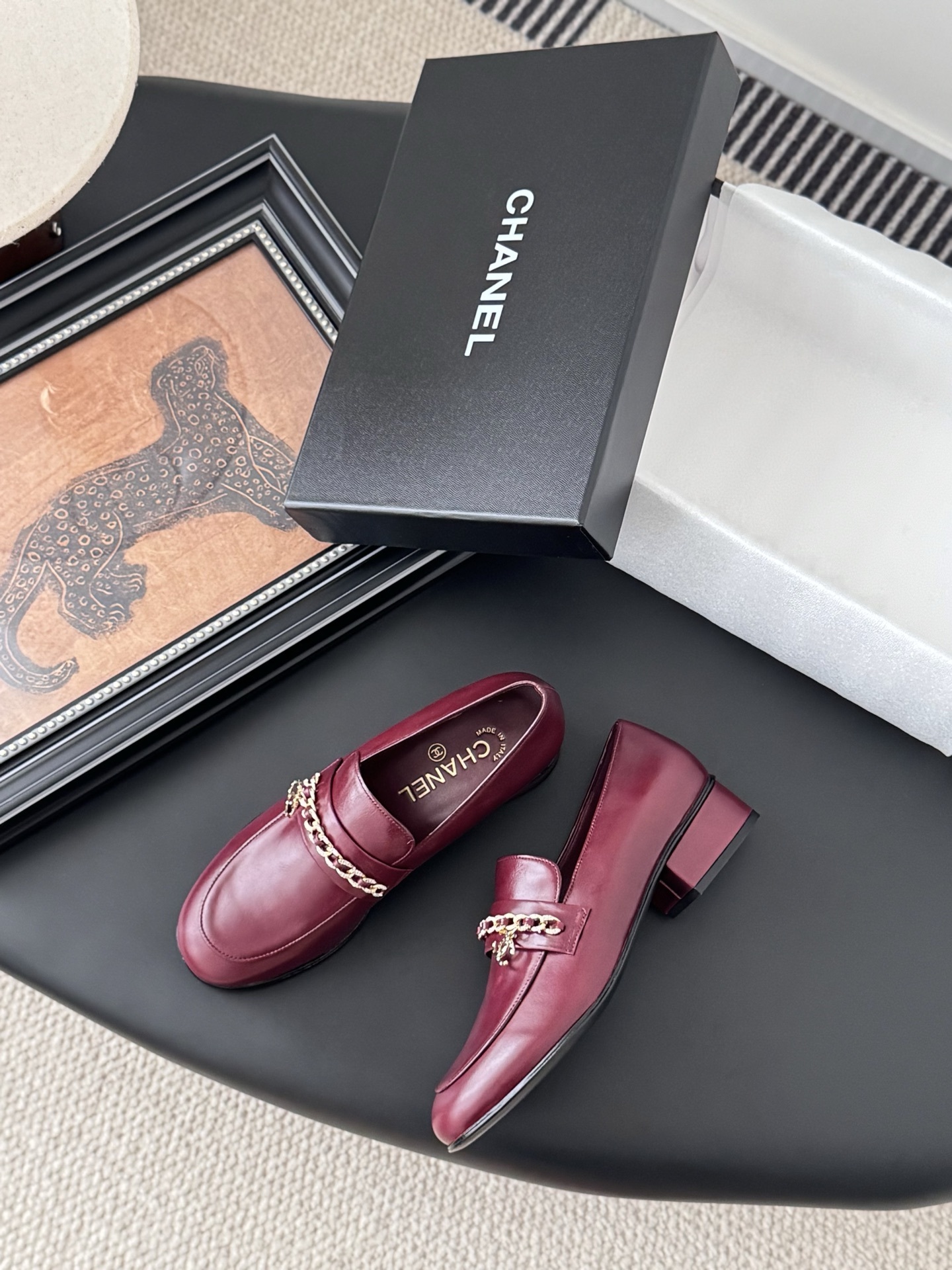 Pair of burgundy Chanel leather loafers with gold chain detail and block heels next to a black Chanel shoe box. - Hình 3