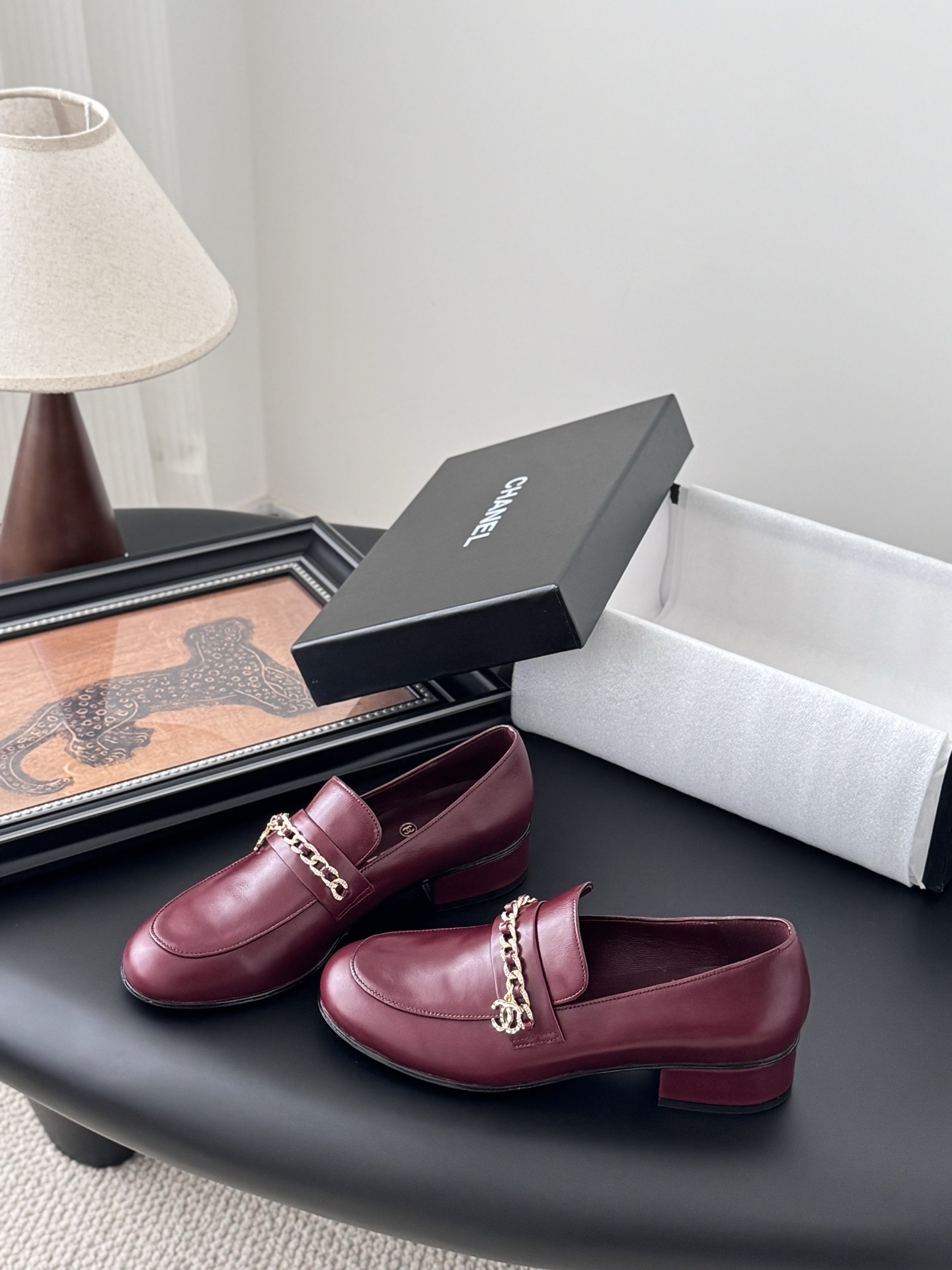 Pair of burgundy Chanel leather loafers with gold chain detail and block heels next to a black Chanel shoe box. - Hình 2