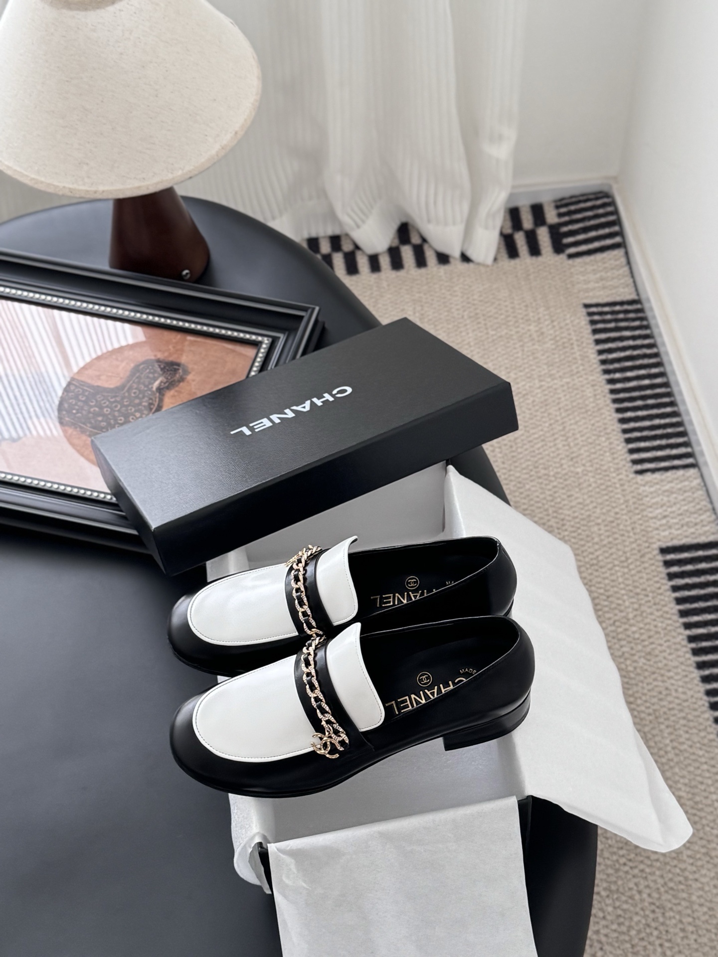 A pair of Chanel black and white leather loafers with gold-tone chain and CC logo sitting next to a Chanel shoe box. - Hình 9