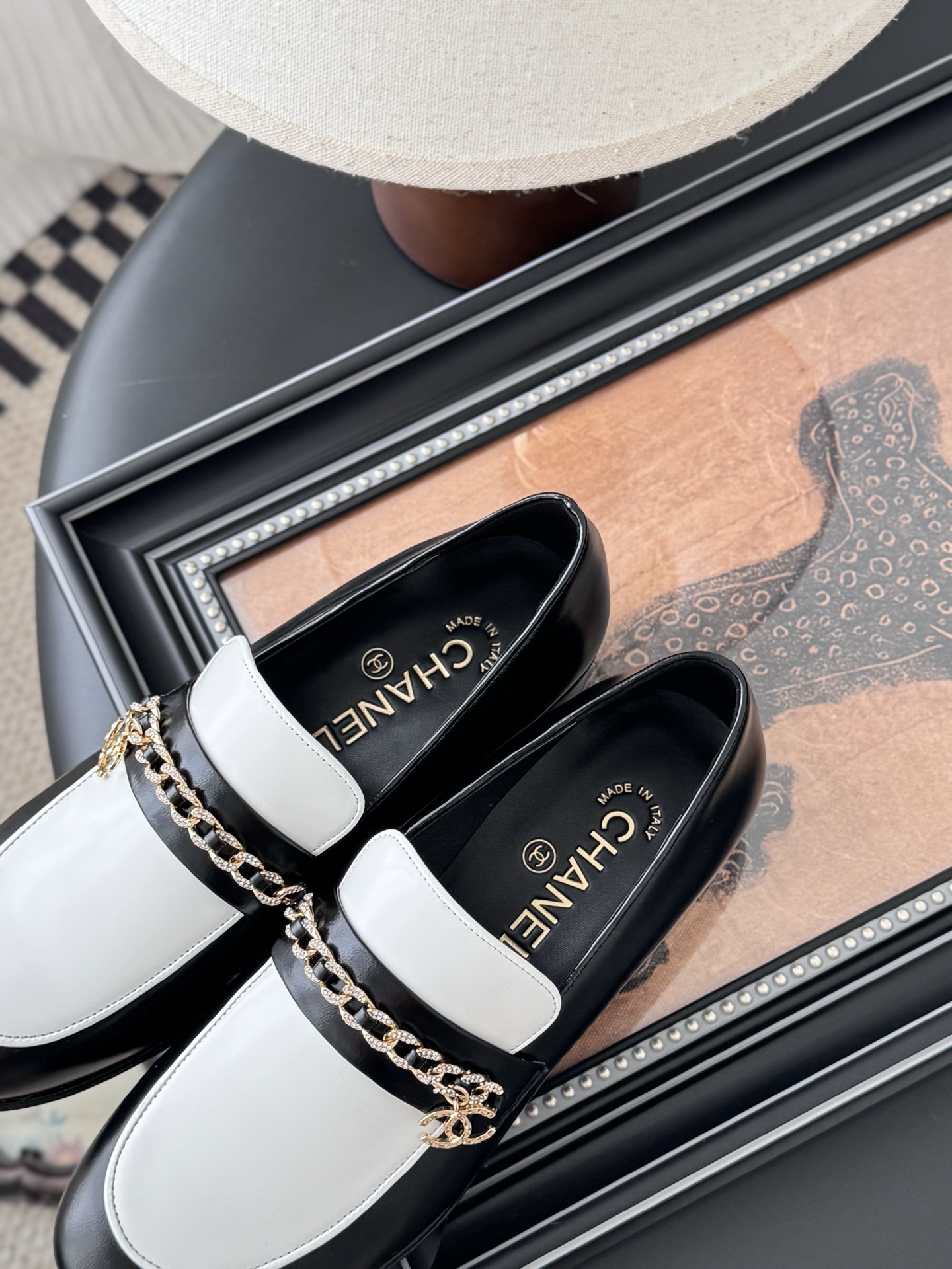 A pair of Chanel black and white leather loafers with gold-tone chain and CC logo sitting next to a Chanel shoe box. - Hình 8