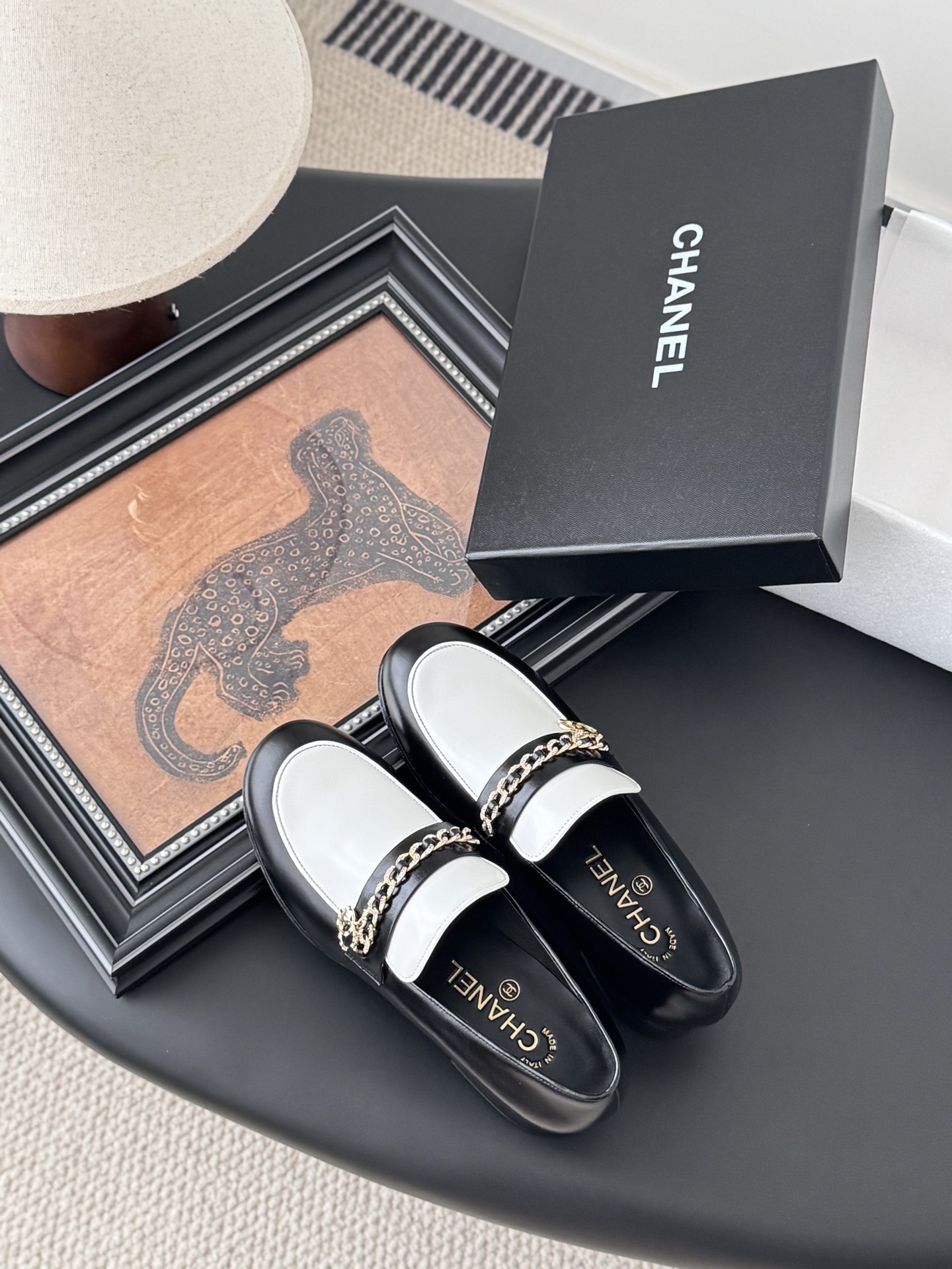 A pair of Chanel black and white leather loafers with gold-tone chain and CC logo sitting next to a Chanel shoe box. - Hình 7