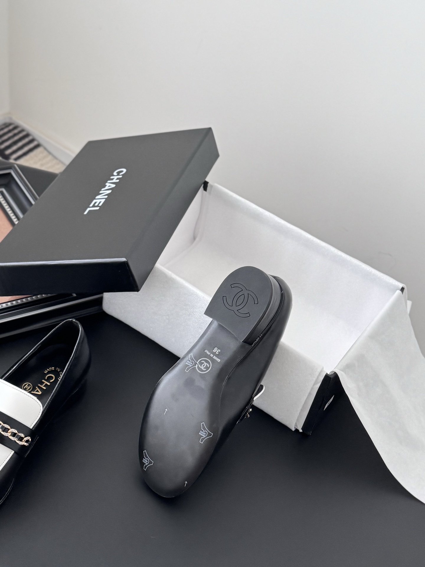 A pair of Chanel black and white leather loafers with gold-tone chain and CC logo sitting next to a Chanel shoe box. - Hình 4