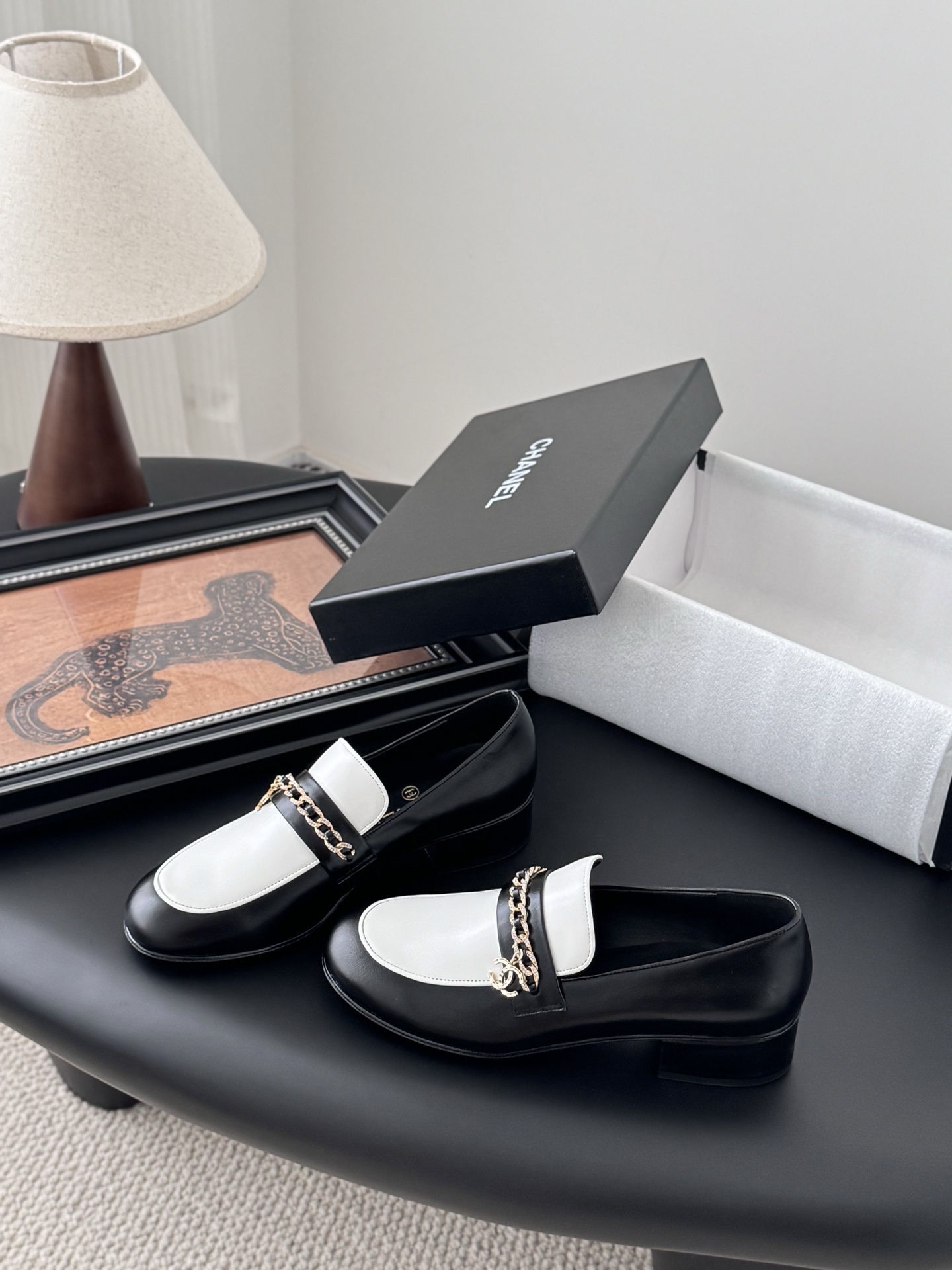 A pair of Chanel black and white leather loafers with gold-tone chain and CC logo sitting next to a Chanel shoe box. - Hình 2