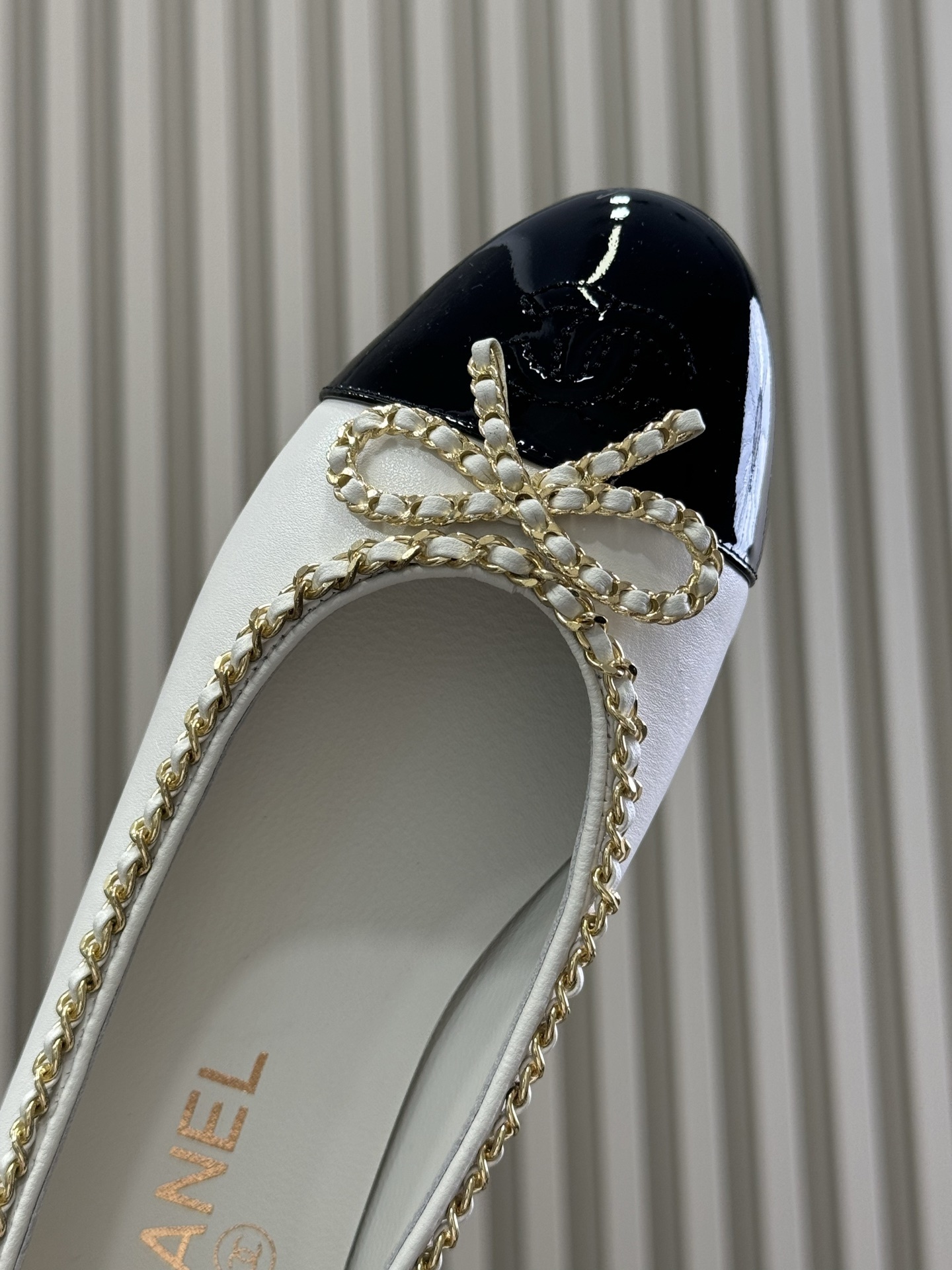A pair of Chanel white leather ballet flats with black patent toe caps and gold chain detailing resting on a black designer box. - Hình 4