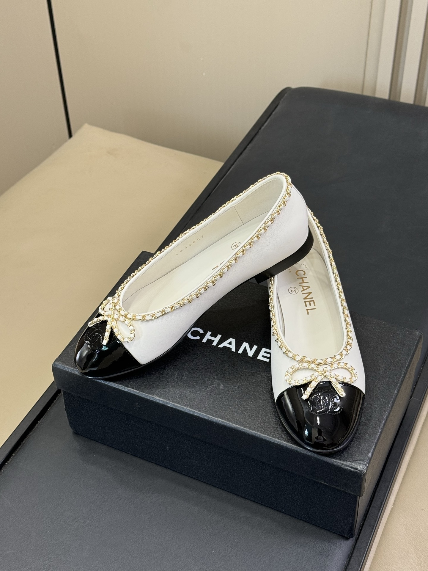 A pair of Chanel white leather ballet flats with black patent toe caps and gold chain detailing resting on a black designer box. - Hình 3