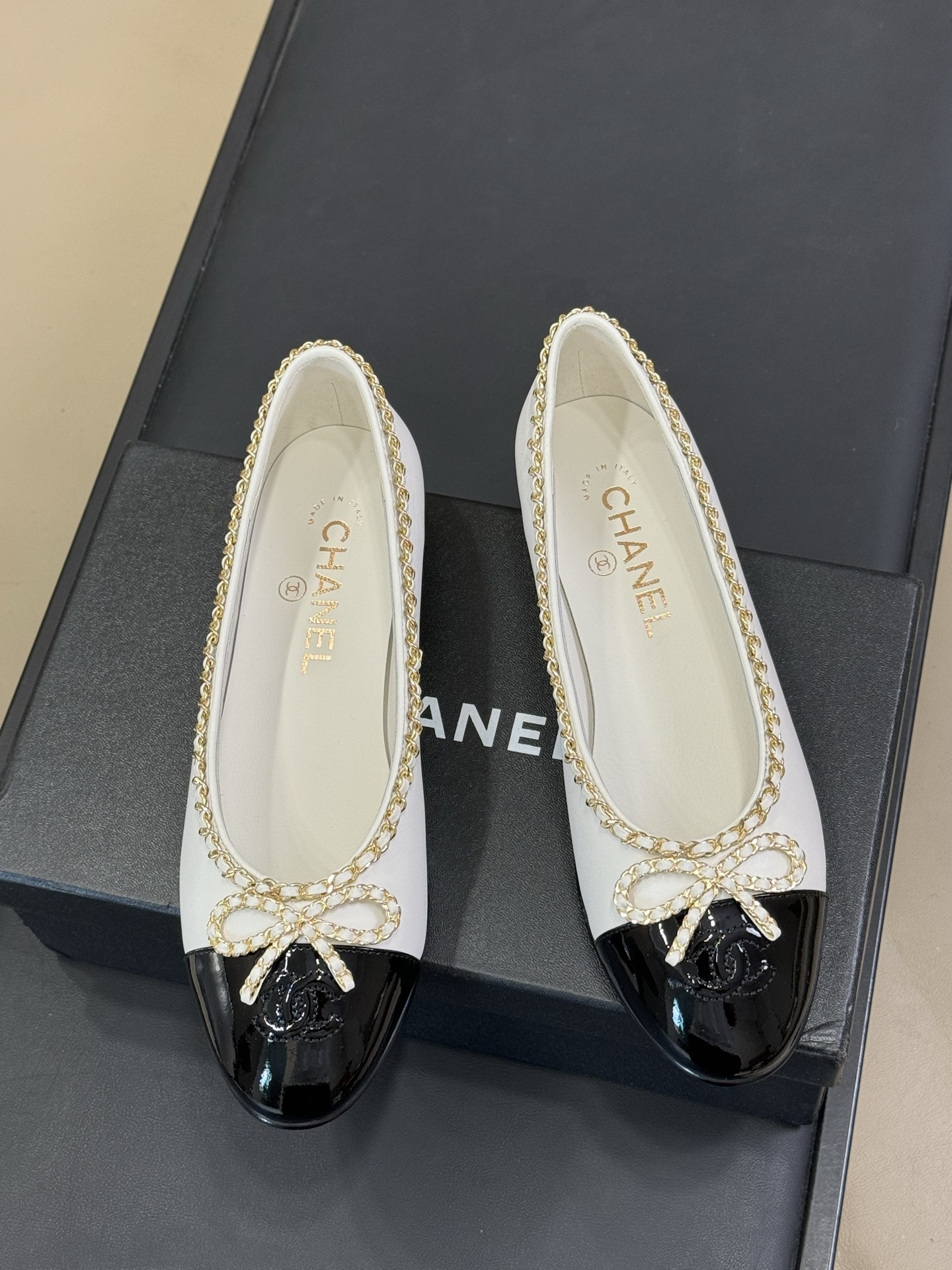 A pair of Chanel white leather ballet flats with black patent toe caps and gold chain detailing resting on a black designer box. - Hình 2
