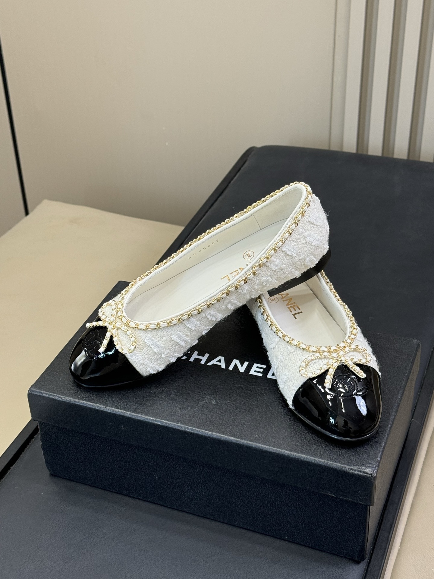 Luxury Chanel ballet flats made of white tweed and black patent leather with gold chain trim and bow detail on a black box. - Hình 3