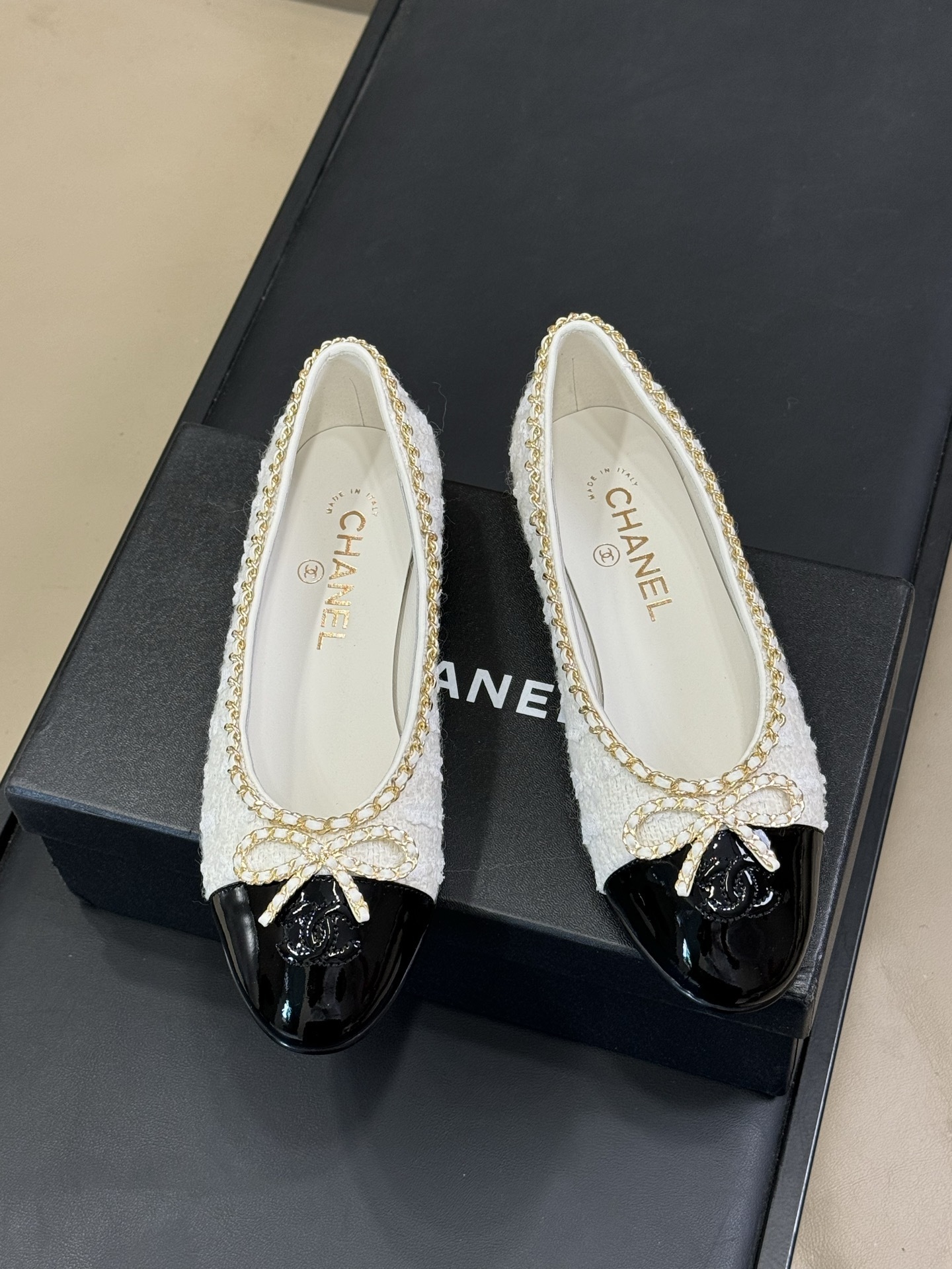 Luxury Chanel ballet flats made of white tweed and black patent leather with gold chain trim and bow detail on a black box. - Hình 2