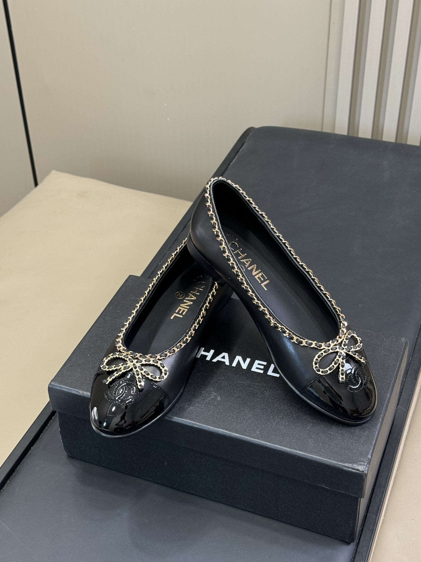 Pair of Chanel black leather ballet flats with patent cap toe and gold chain trim on a black box - Hình 3