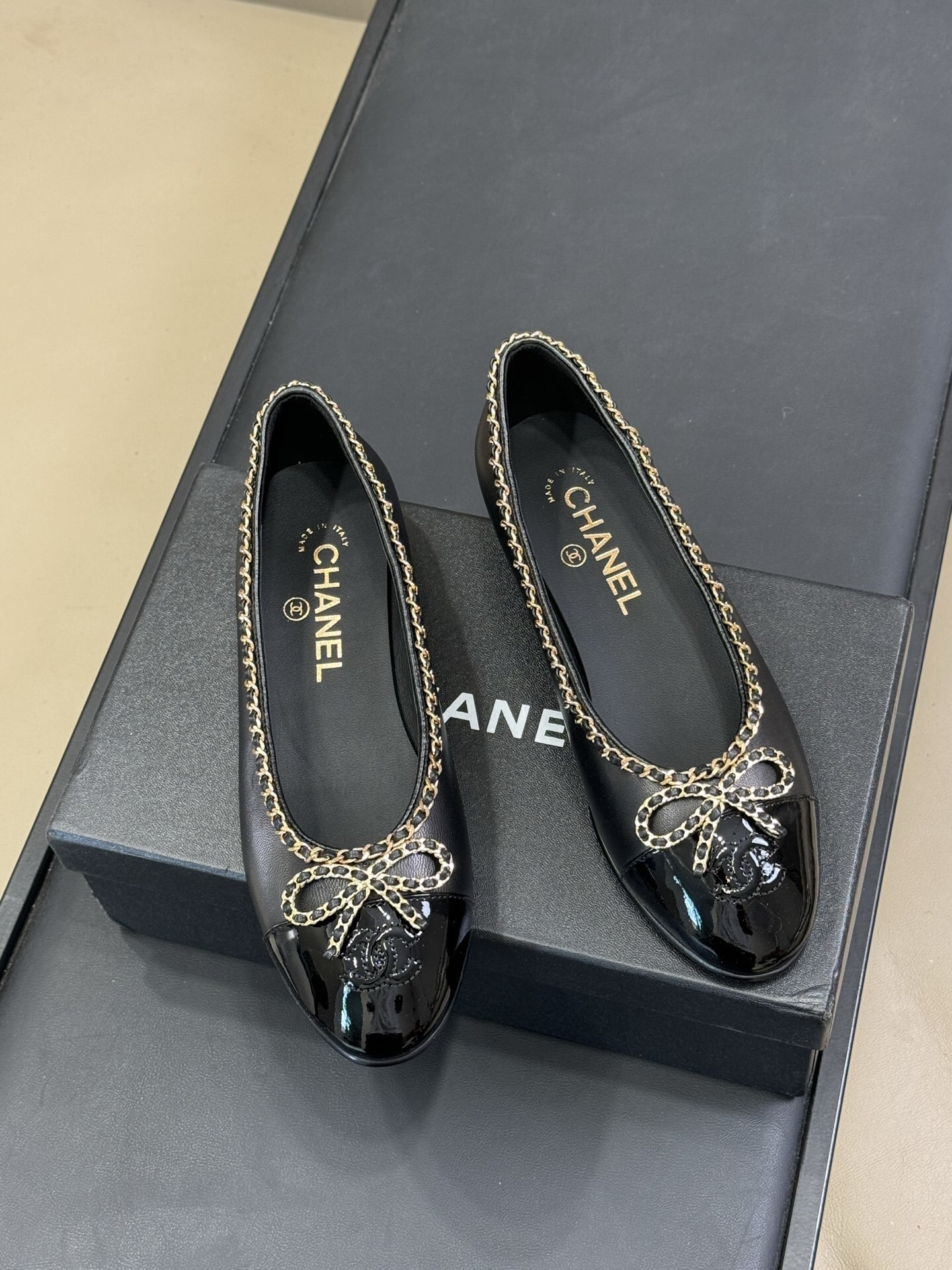 Pair of Chanel black leather ballet flats with patent cap toe and gold chain trim on a black box - Hình 2