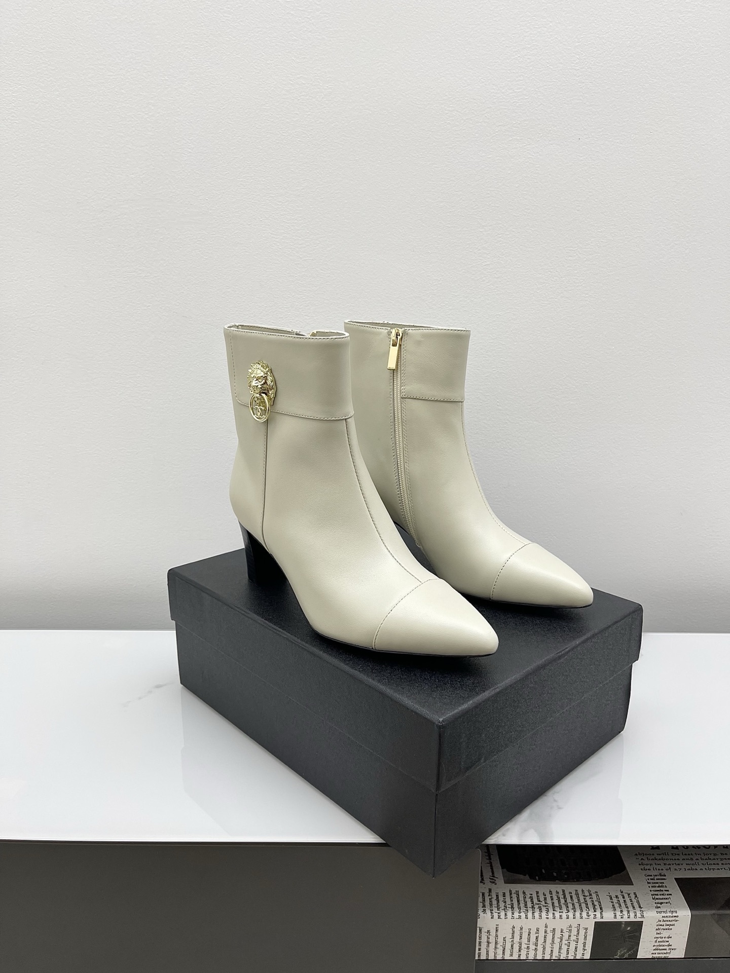 Pair of Chanel ivory leather pointed-toe ankle boots with gold lion head emblem and side zipper on a white marble surface. - Hình 7
