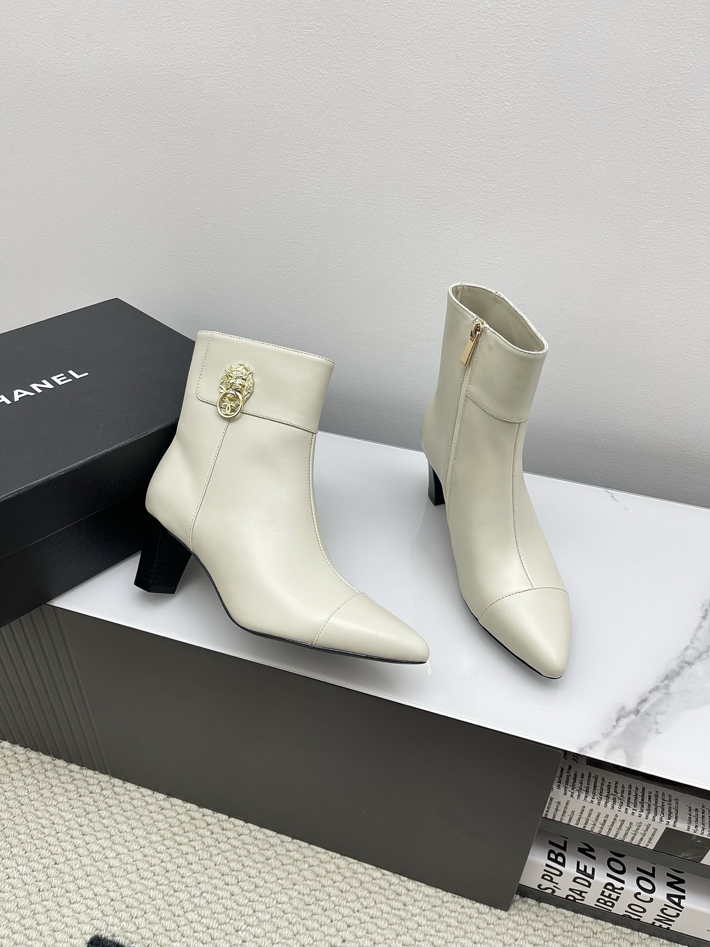 Pair of Chanel ivory leather pointed-toe ankle boots with gold lion head emblem and side zipper on a white marble surface. - Hình 5