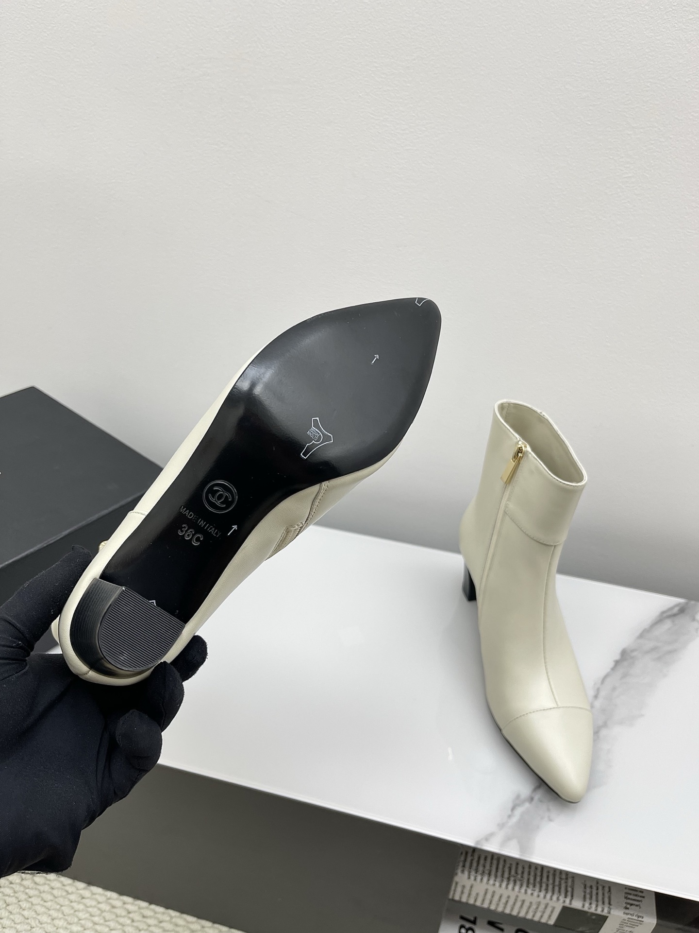 Pair of Chanel ivory leather pointed-toe ankle boots with gold lion head emblem and side zipper on a white marble surface. - Hình 4