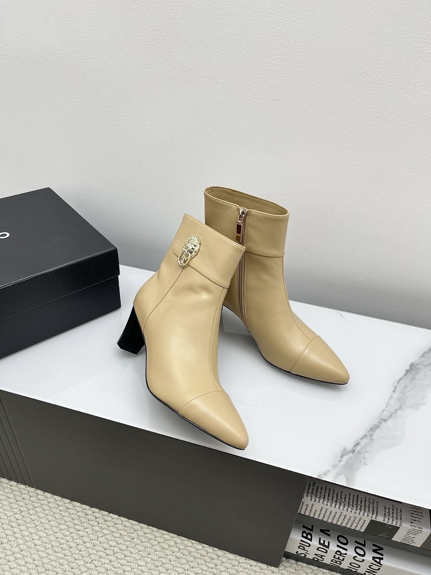 A pair of elegant beige leather pointed-toe ankle boots with a golden lion emblem and black block heel displayed on a white surface. - Hình 3