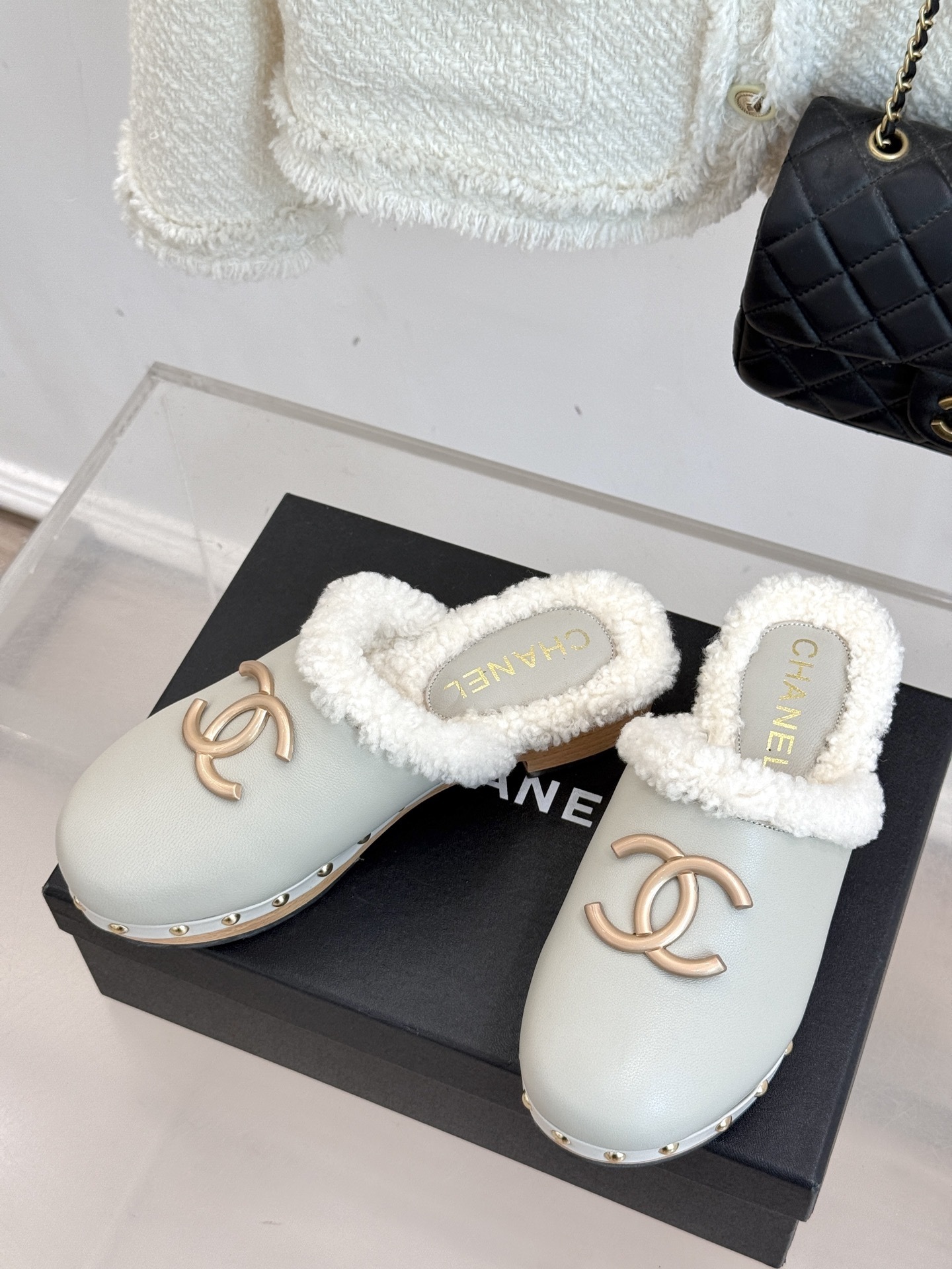 Pair of luxury off-white Chanel leather clogs with shearling lining and gold-tone CC logo displayed on a black box. - Hình 2