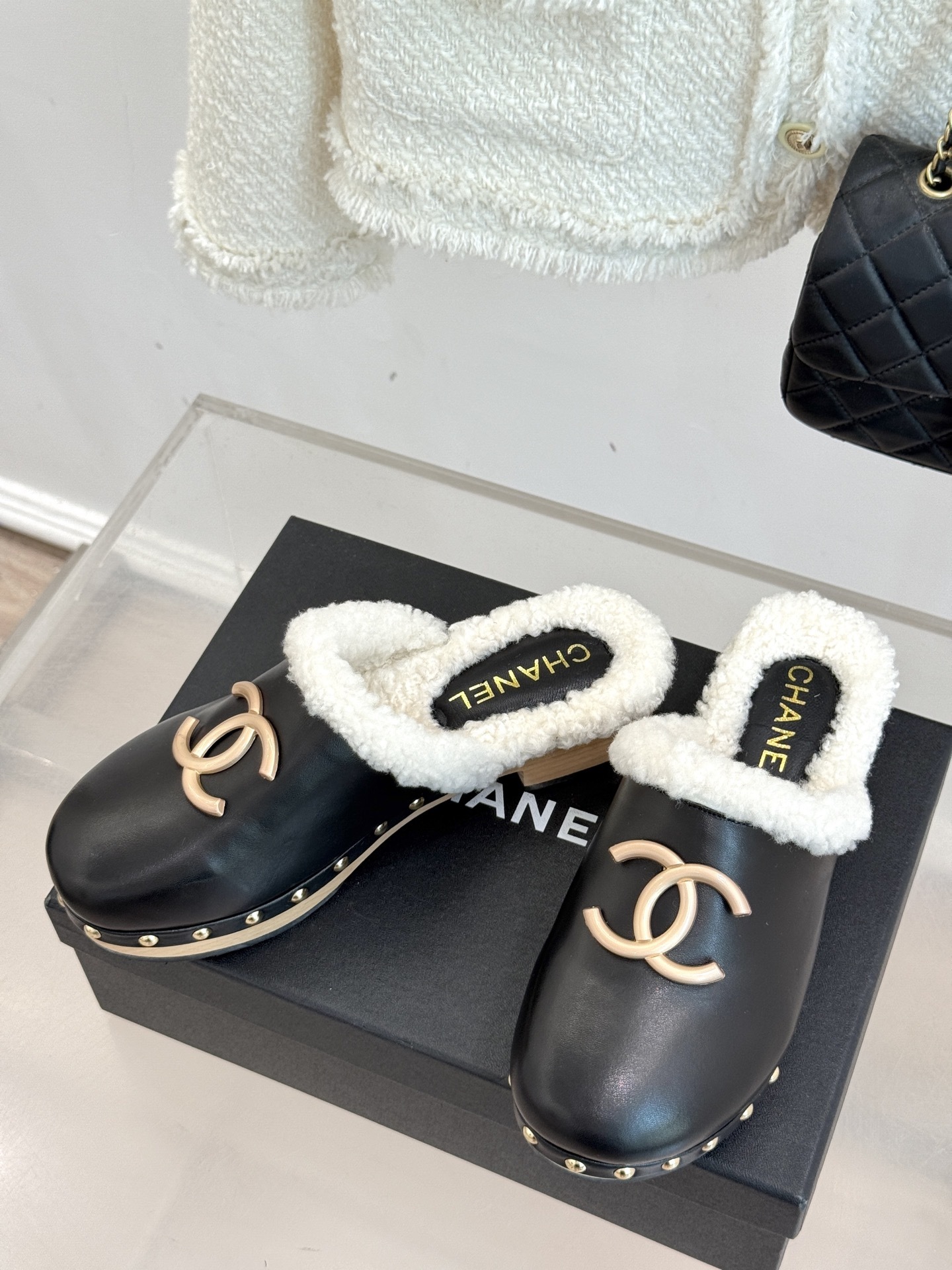 Pair of Chanel black leather clogs with white shearling lining, gold CC logo, and studded wooden soles resting on a black box - Hình 2