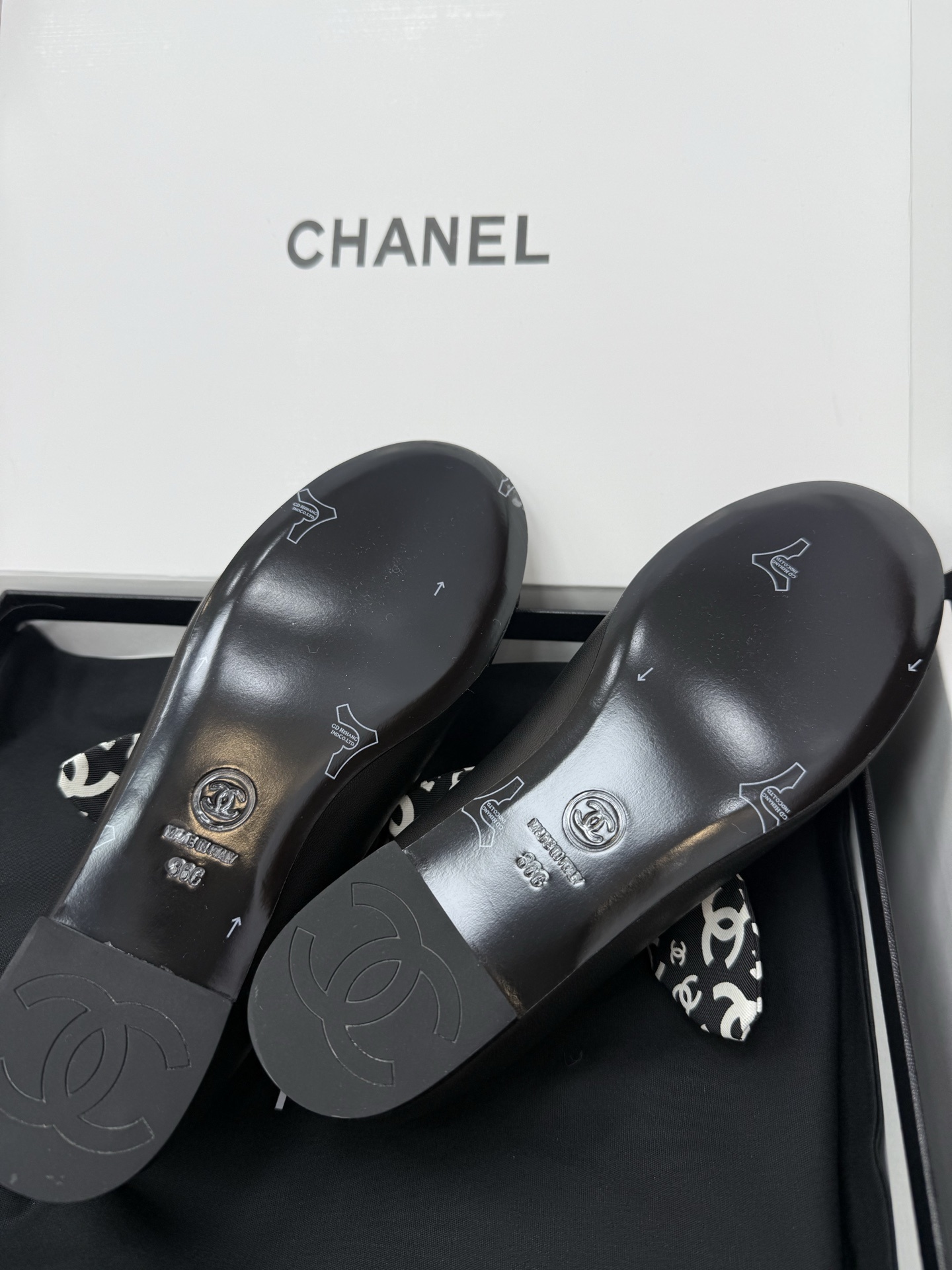 A pair of black Chanel leather ballet flats featuring a fabric bow with white CC logos and a quilted insole, resting on a black box. - Hình 9