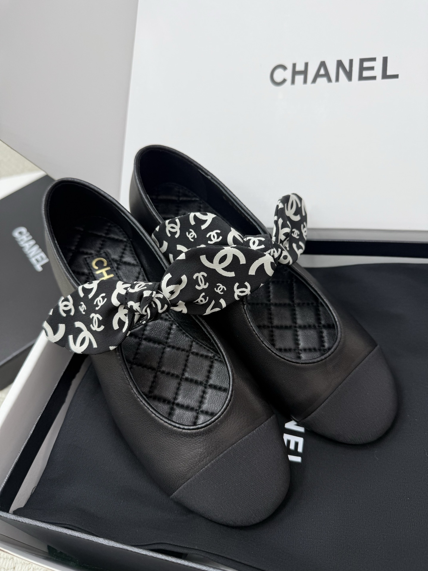 A pair of black Chanel leather ballet flats featuring a fabric bow with white CC logos and a quilted insole, resting on a black box. - Hình 7