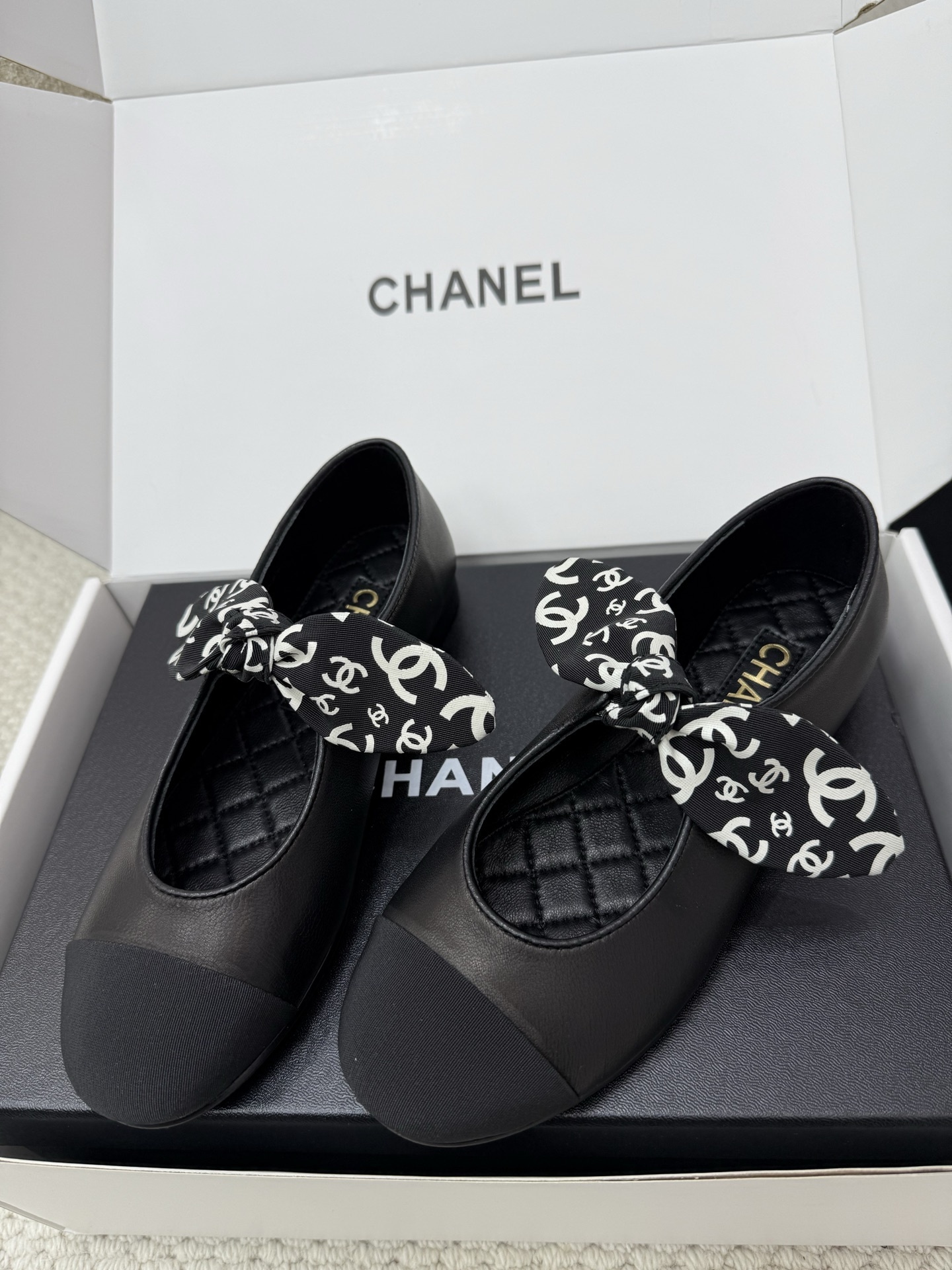 A pair of black Chanel leather ballet flats featuring a fabric bow with white CC logos and a quilted insole, resting on a black box. - Hình 5