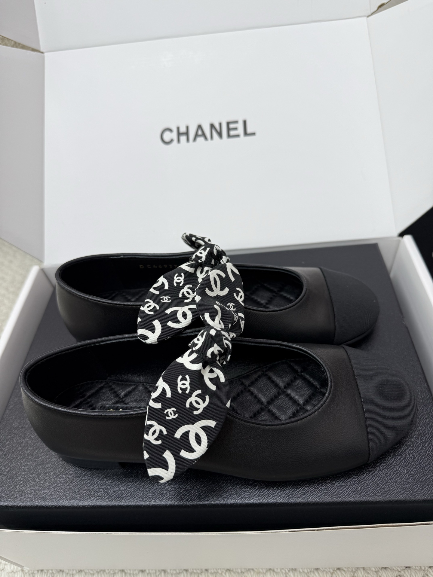 A pair of black Chanel leather ballet flats featuring a fabric bow with white CC logos and a quilted insole, resting on a black box. - Hình 4