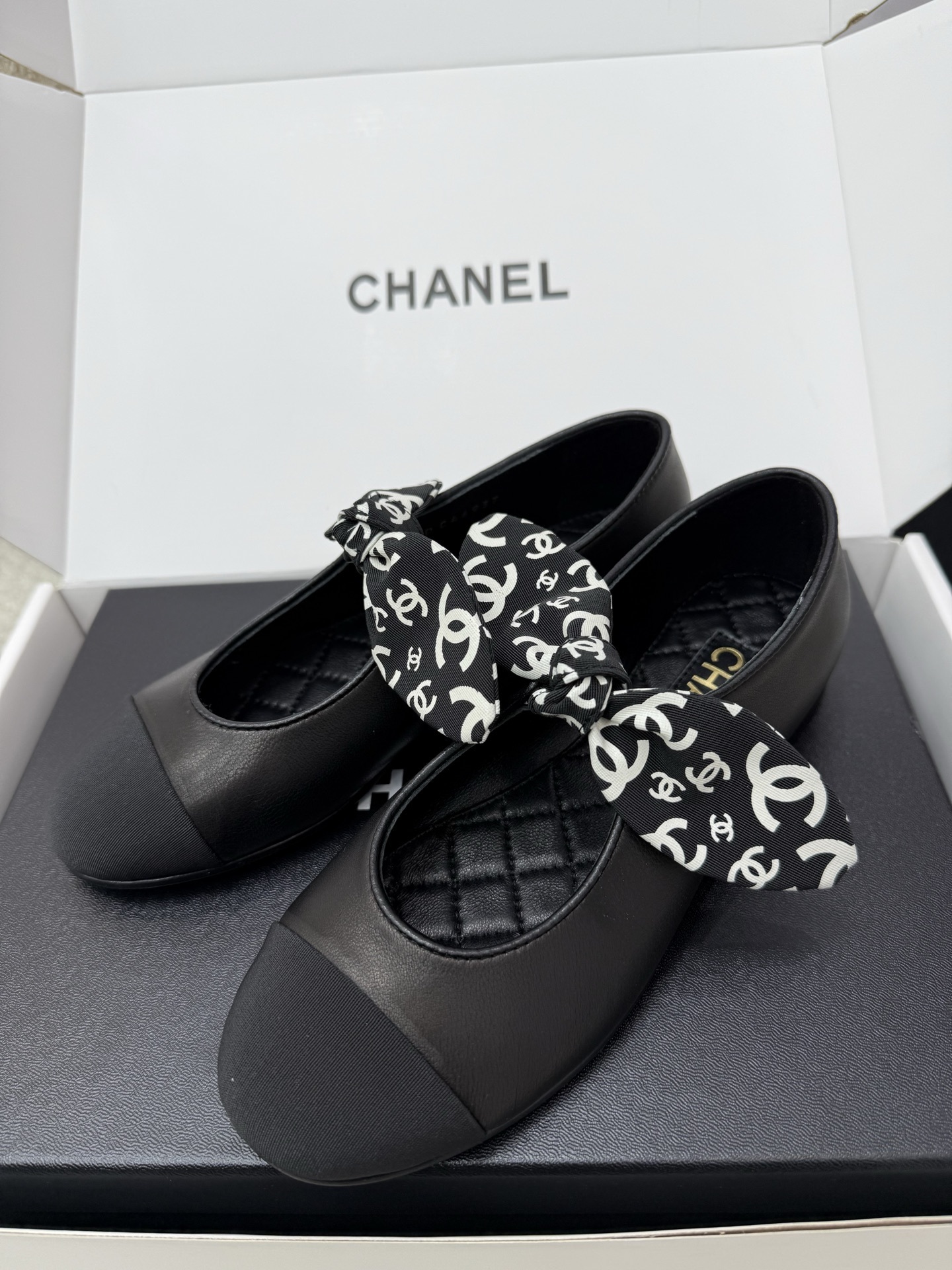 A pair of black Chanel leather ballet flats featuring a fabric bow with white CC logos and a quilted insole, resting on a black box. - Hình 3