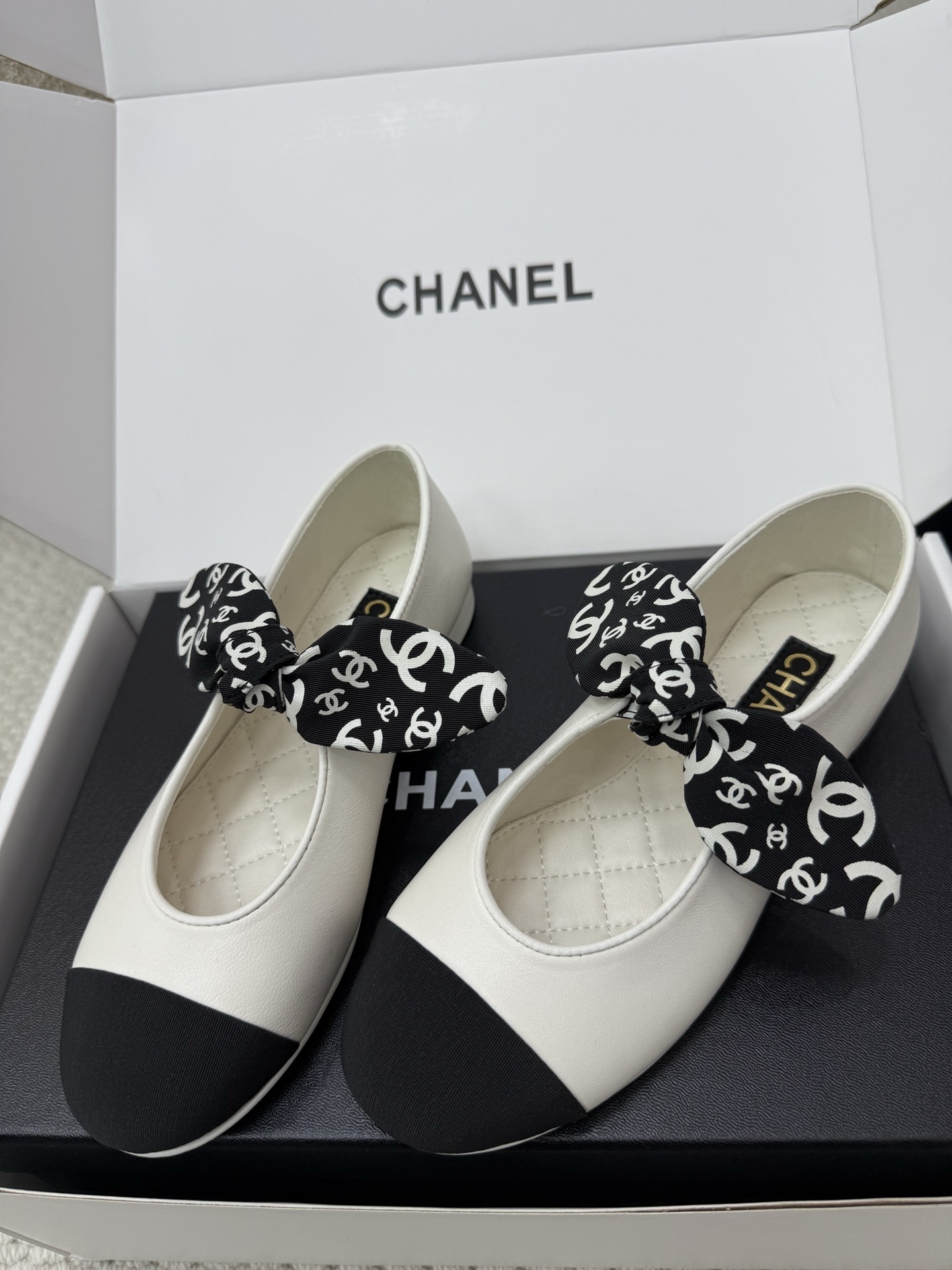 A pair of Chanel white leather ballet flats with black toe caps and a decorative black and white CC logo ribbon bow, presented in a Chanel box. - Hình 6