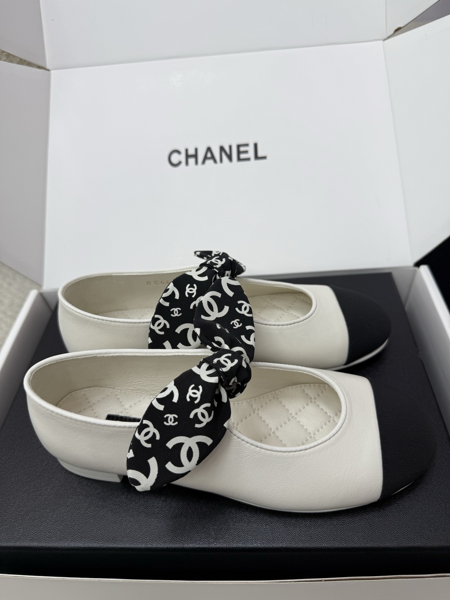 A pair of Chanel white leather ballet flats with black toe caps and a decorative black and white CC logo ribbon bow, presented in a Chanel box. - Hình 4