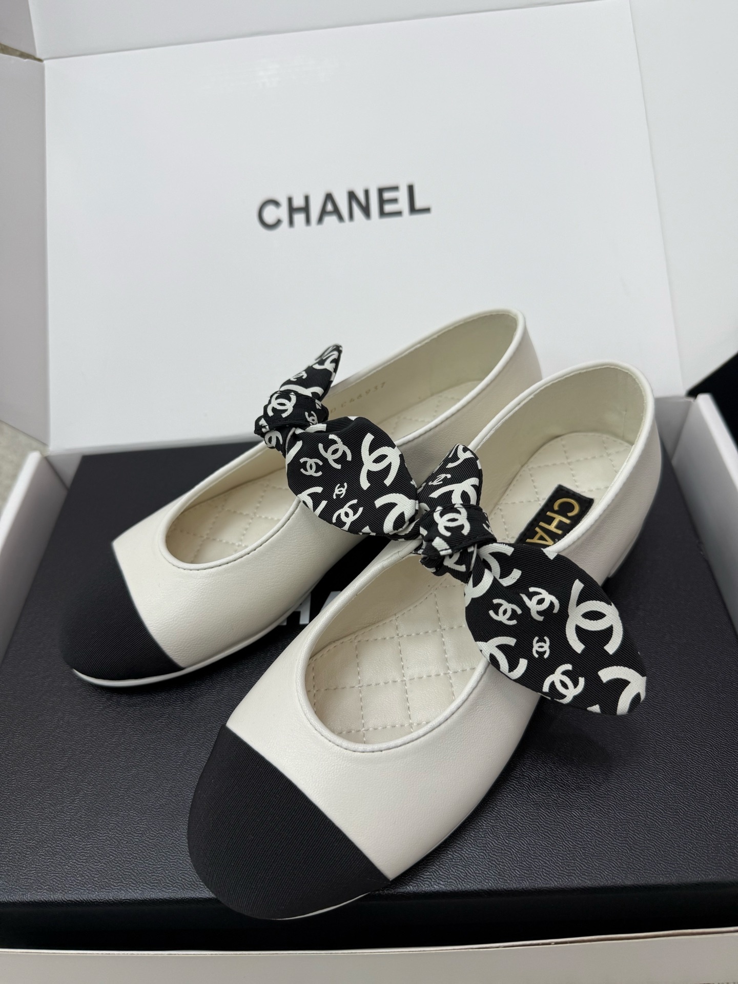 A pair of Chanel white leather ballet flats with black toe caps and a decorative black and white CC logo ribbon bow, presented in a Chanel box. - Hình 3