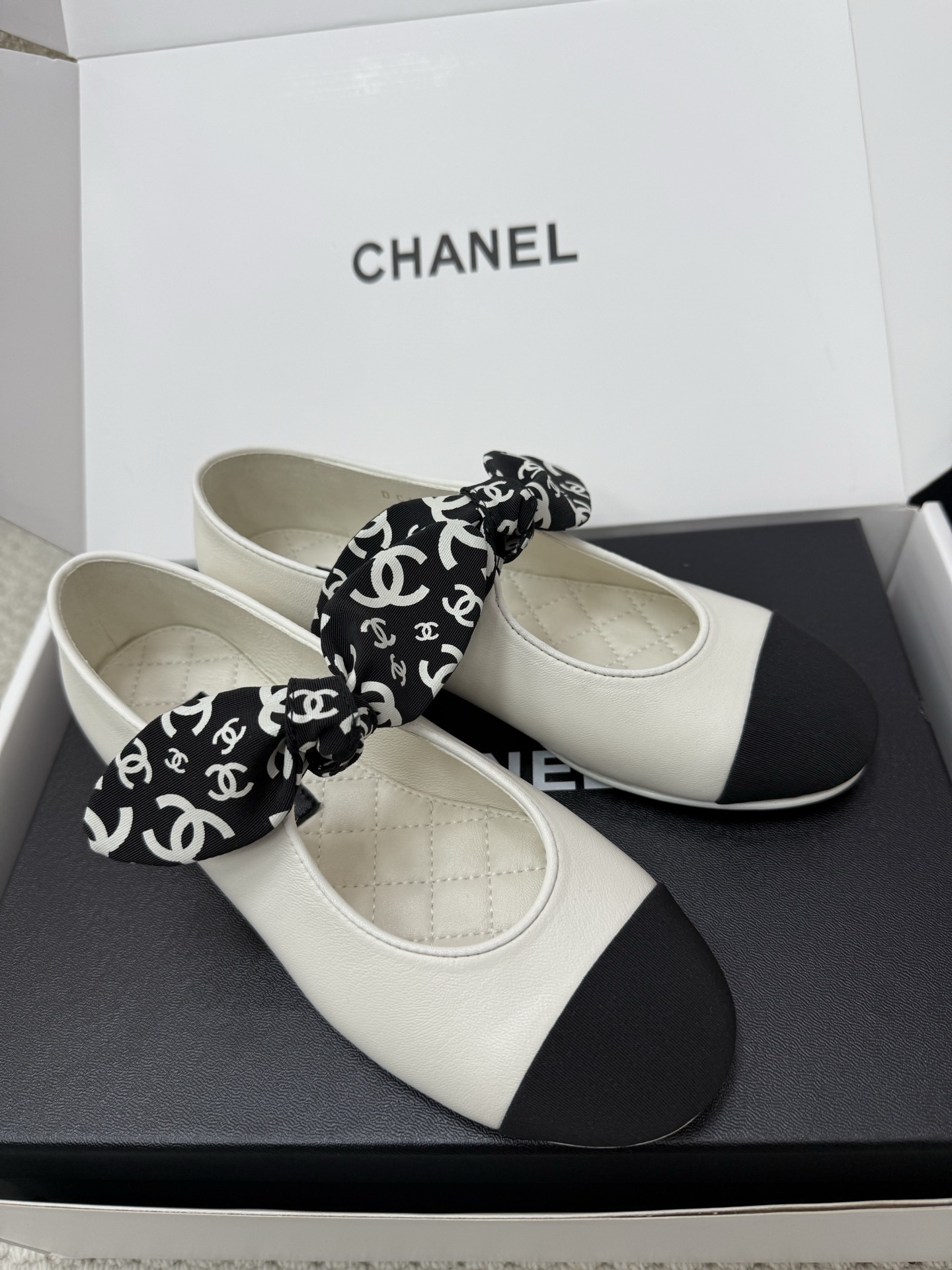A pair of Chanel white leather ballet flats with black toe caps and a decorative black and white CC logo ribbon bow, presented in a Chanel box. - Hình 2
