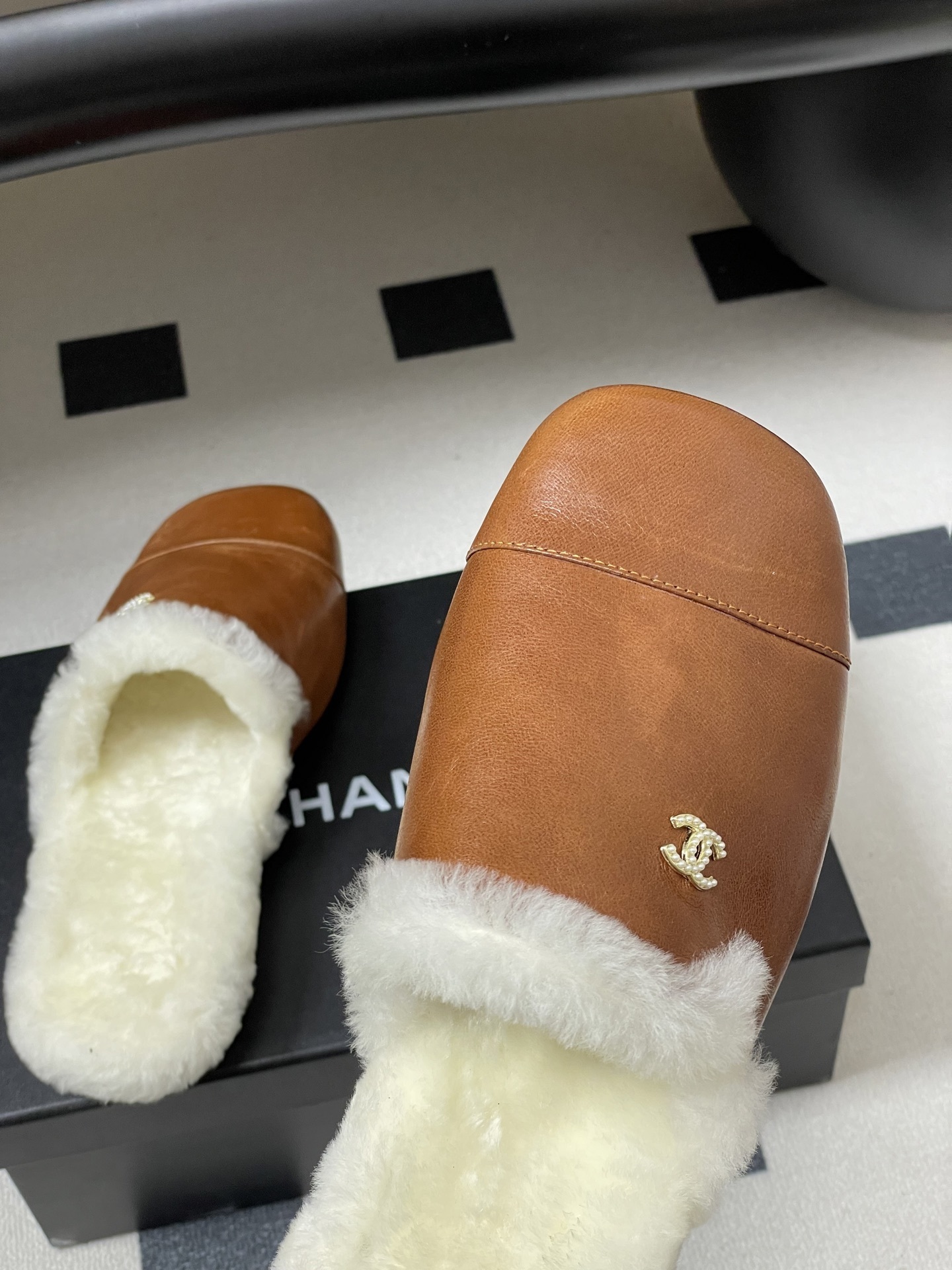 A pair of brown leather slippers with white shearling lining and a gold interlocking C logo on a black surface. - Hình 9
