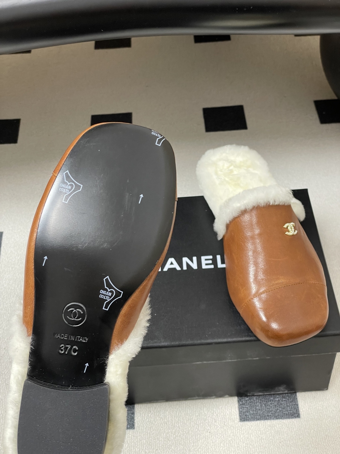 A pair of brown leather slippers with white shearling lining and a gold interlocking C logo on a black surface. - Hình 8