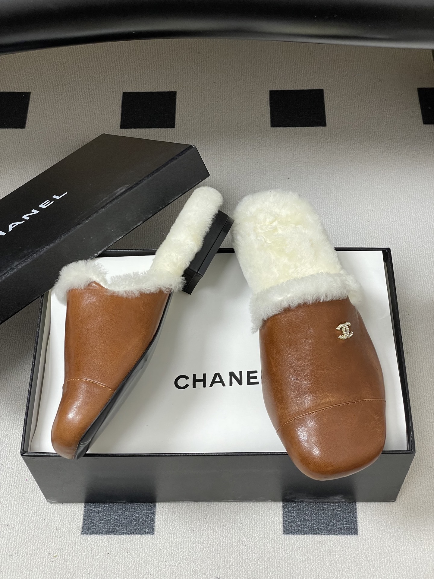 A pair of brown leather slippers with white shearling lining and a gold interlocking C logo on a black surface. - Hình 6