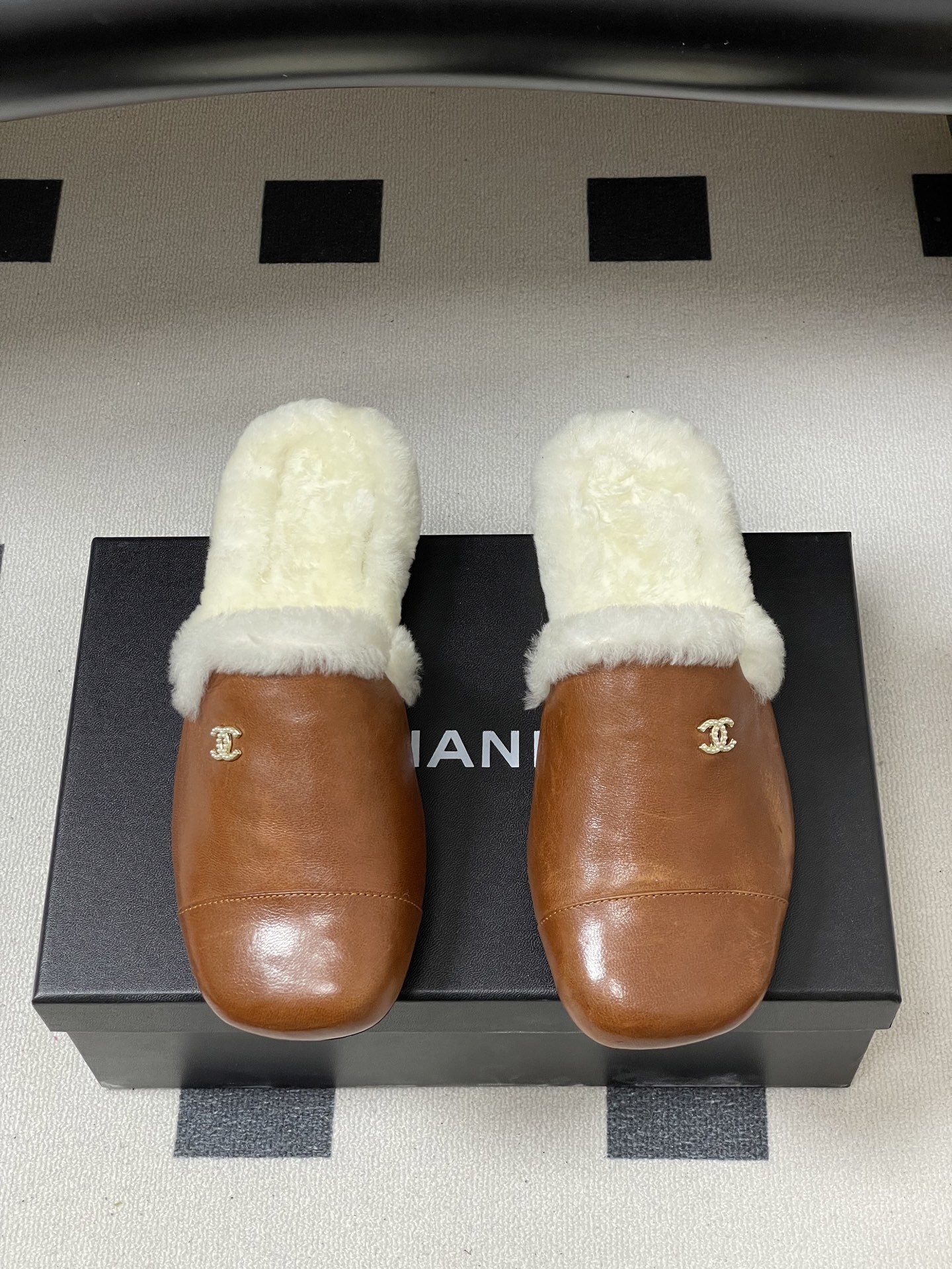 A pair of brown leather slippers with white shearling lining and a gold interlocking C logo on a black surface. - Hình 5