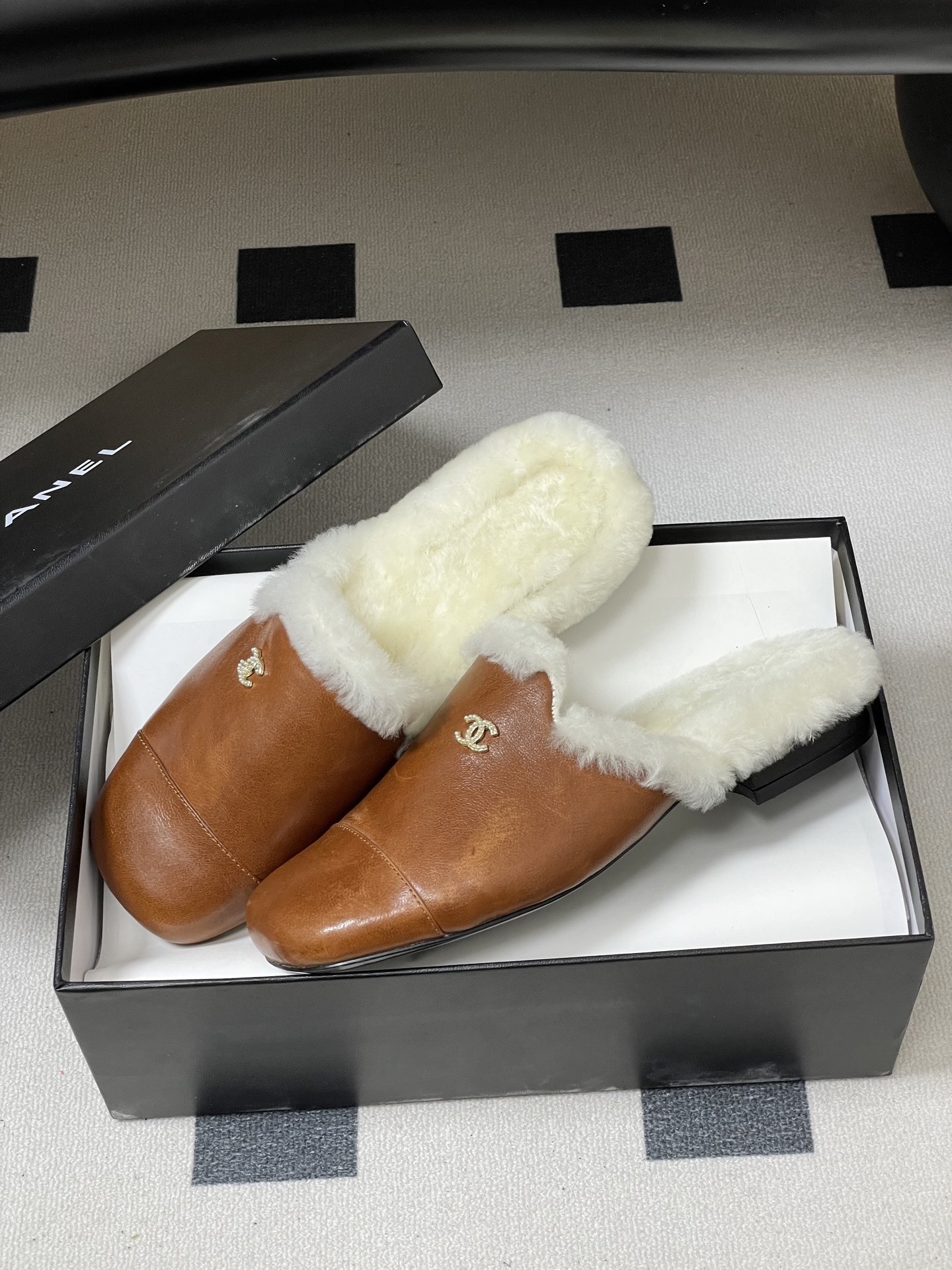 A pair of brown leather slippers with white shearling lining and a gold interlocking C logo on a black surface. - Hình 4