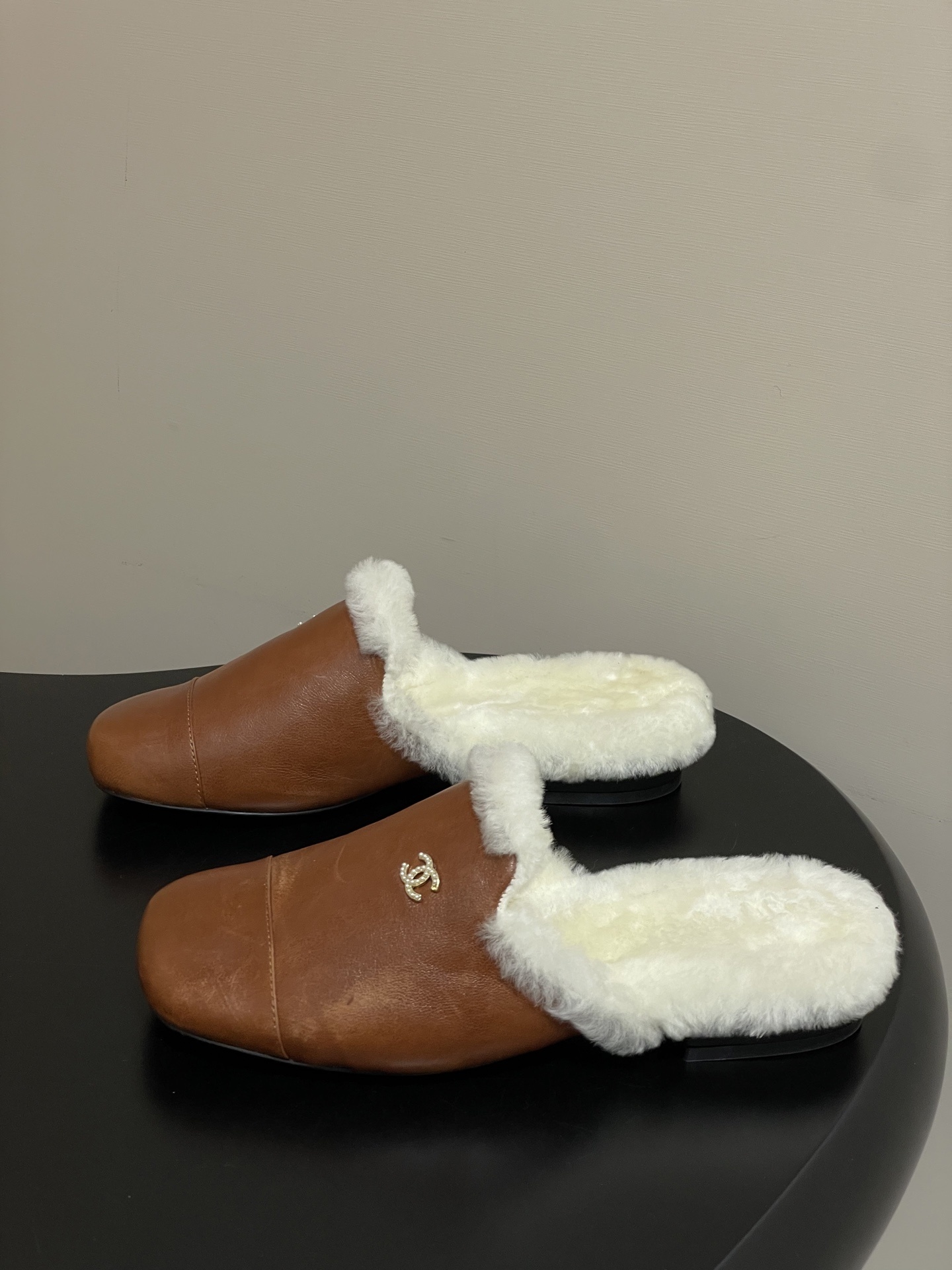 A pair of brown leather slippers with white shearling lining and a gold interlocking C logo on a black surface. - Hình 3