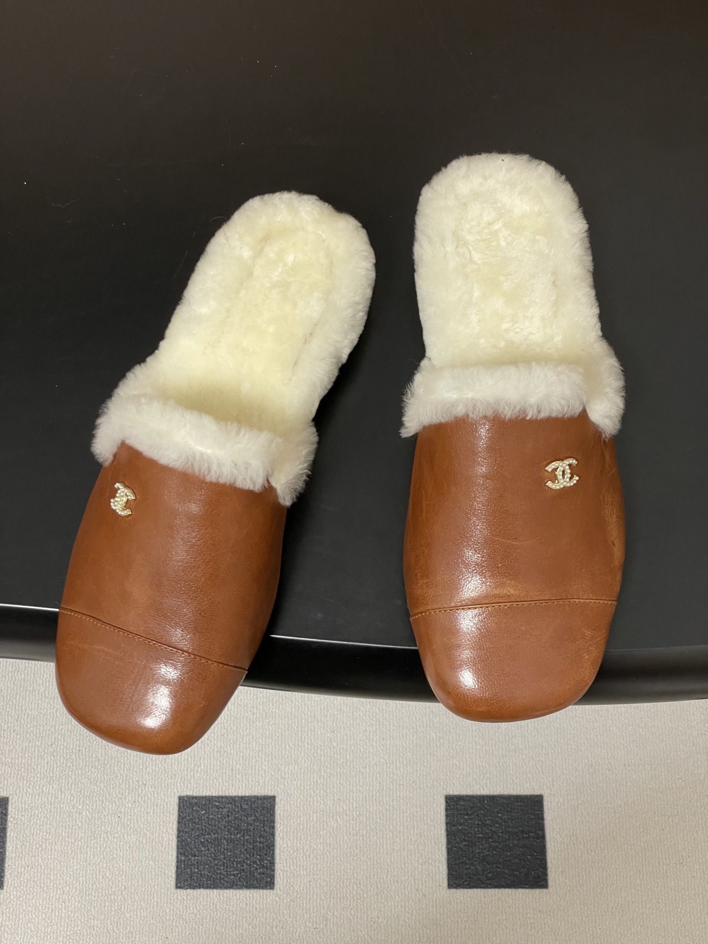 A pair of brown leather slippers with white shearling lining and a gold interlocking C logo on a black surface. - Hình 2