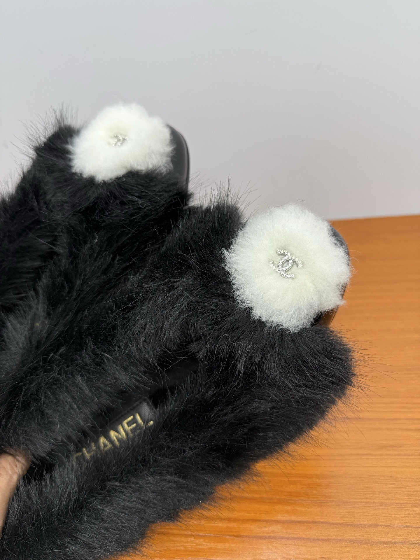 Pair of black faux fur loafers with white pom-poms and crystal CC logos on a wooden table - Hình 7