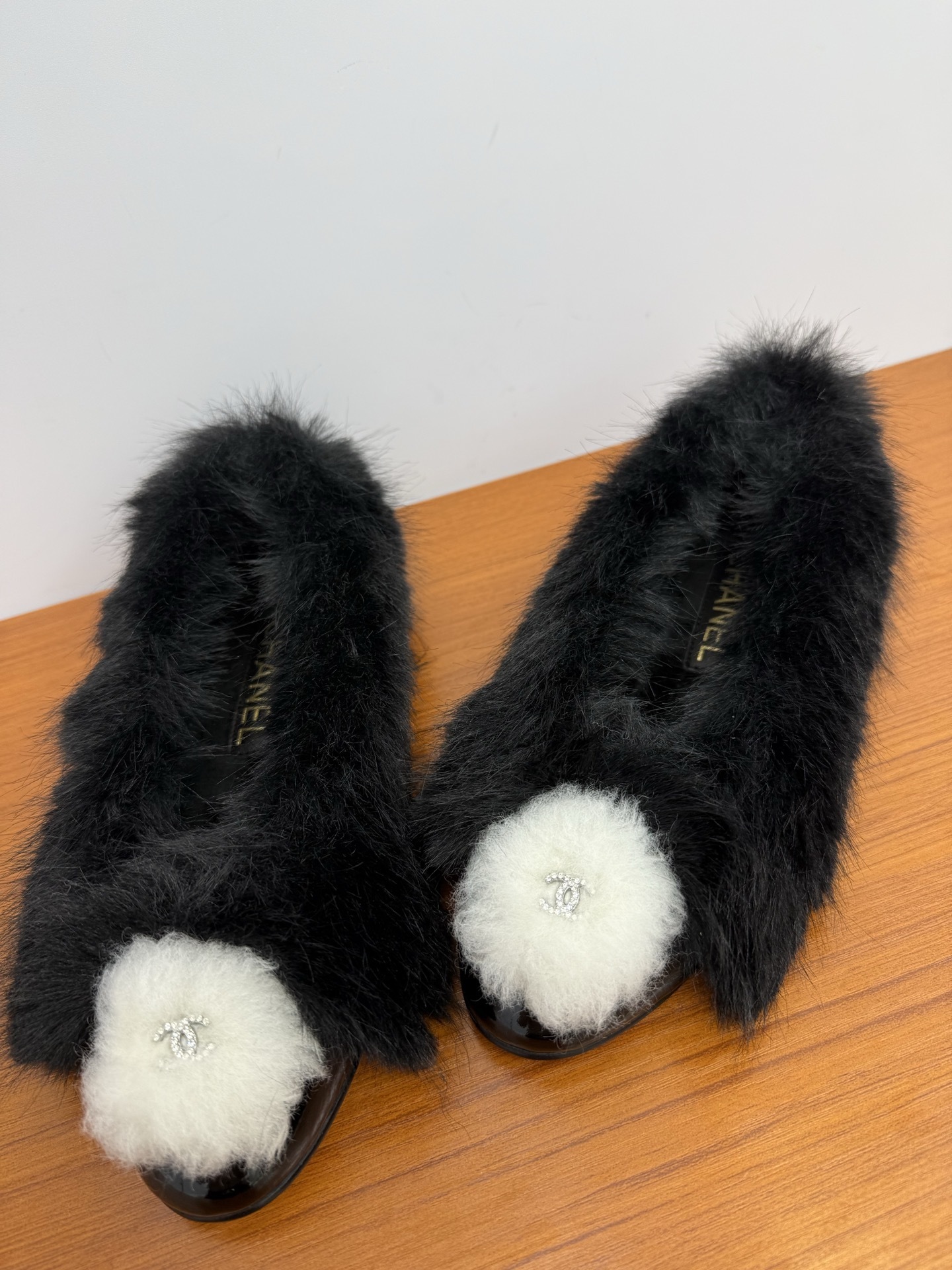 Pair of black faux fur loafers with white pom-poms and crystal CC logos on a wooden table - Hình 6