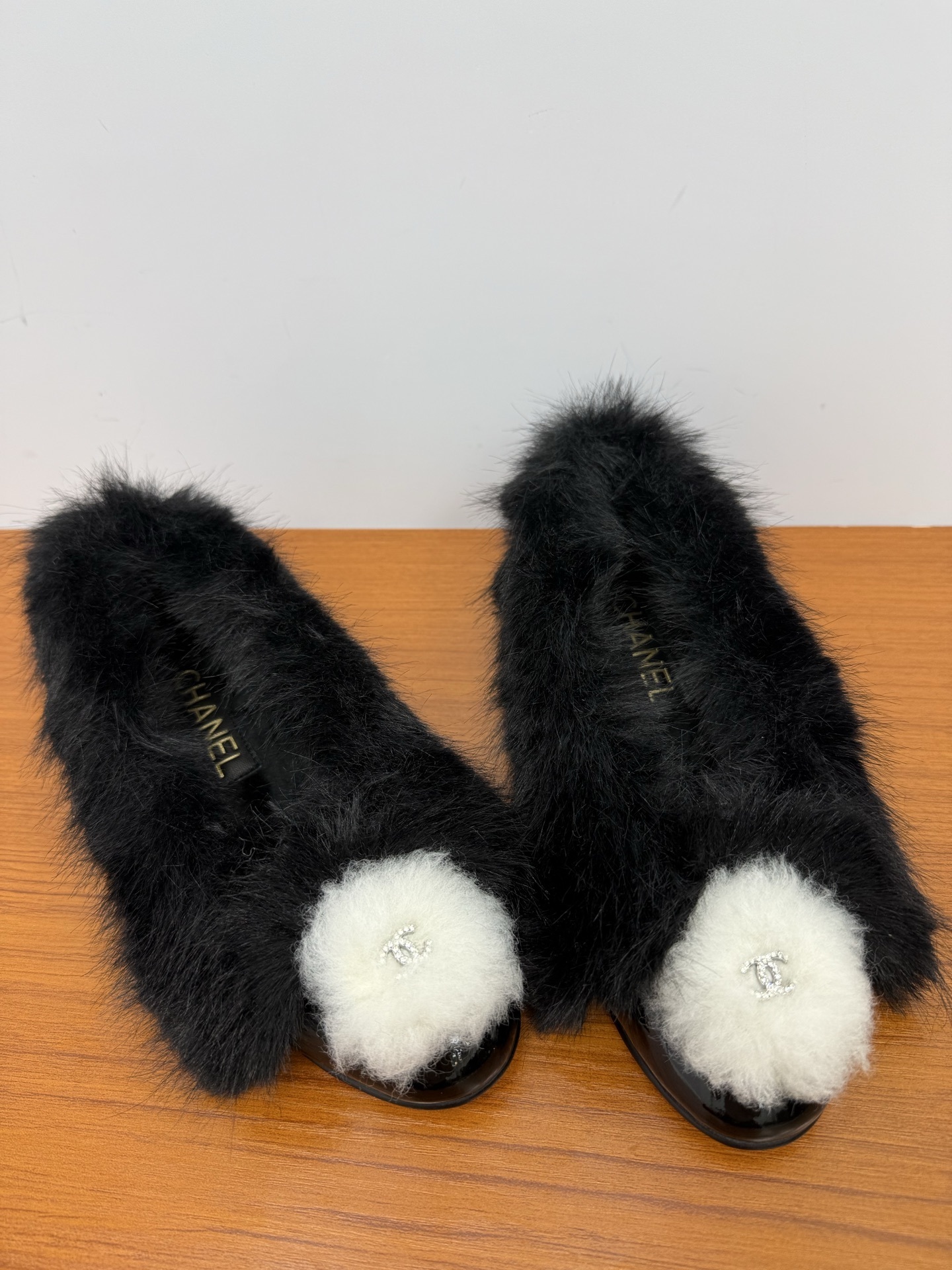 Pair of black faux fur loafers with white pom-poms and crystal CC logos on a wooden table - Hình 5