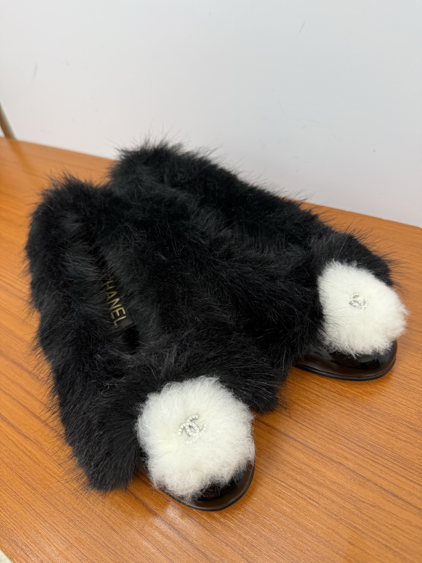 Pair of black faux fur loafers with white pom-poms and crystal CC logos on a wooden table - Hình 4