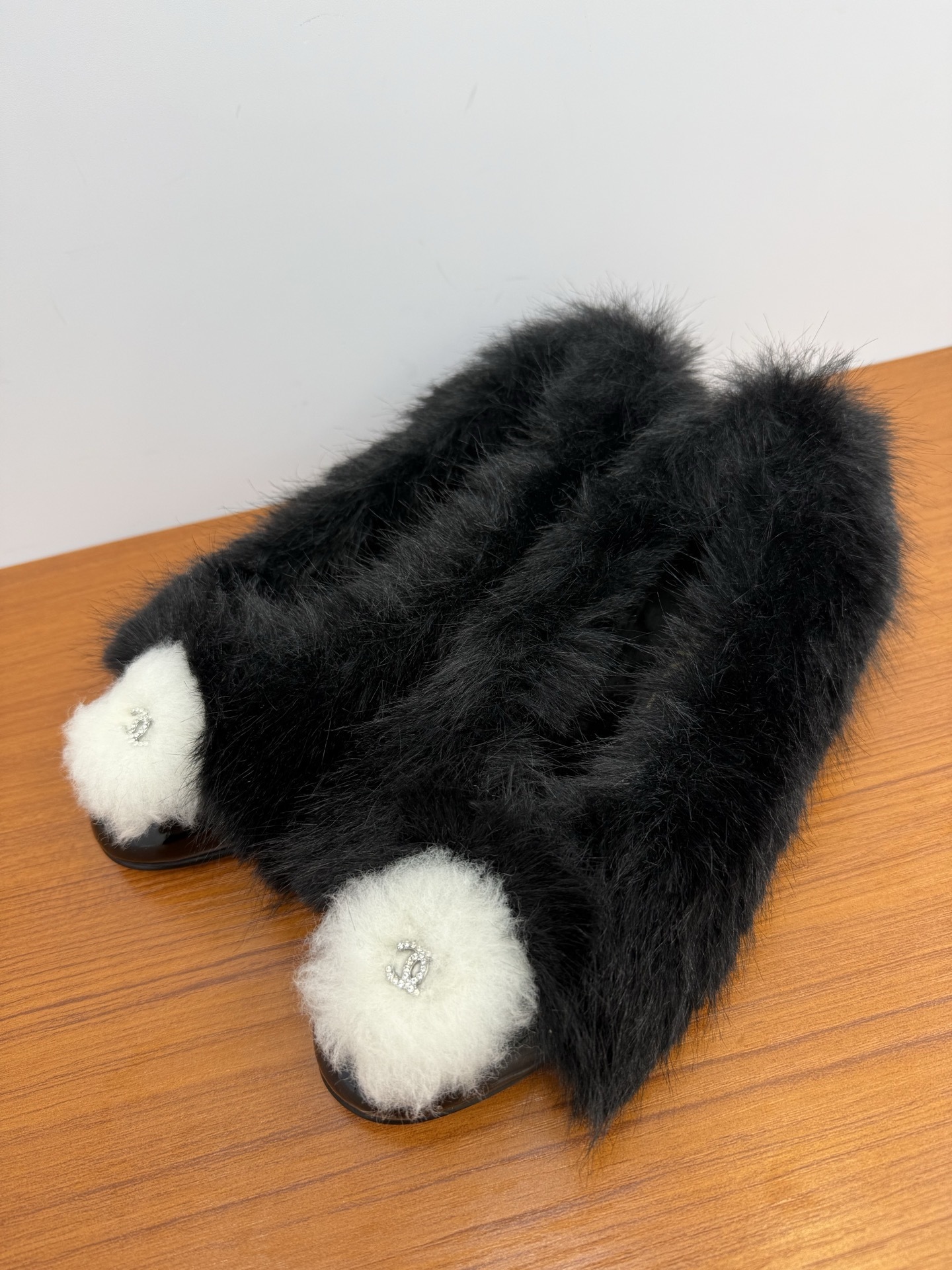 Pair of black faux fur loafers with white pom-poms and crystal CC logos on a wooden table - Hình 3