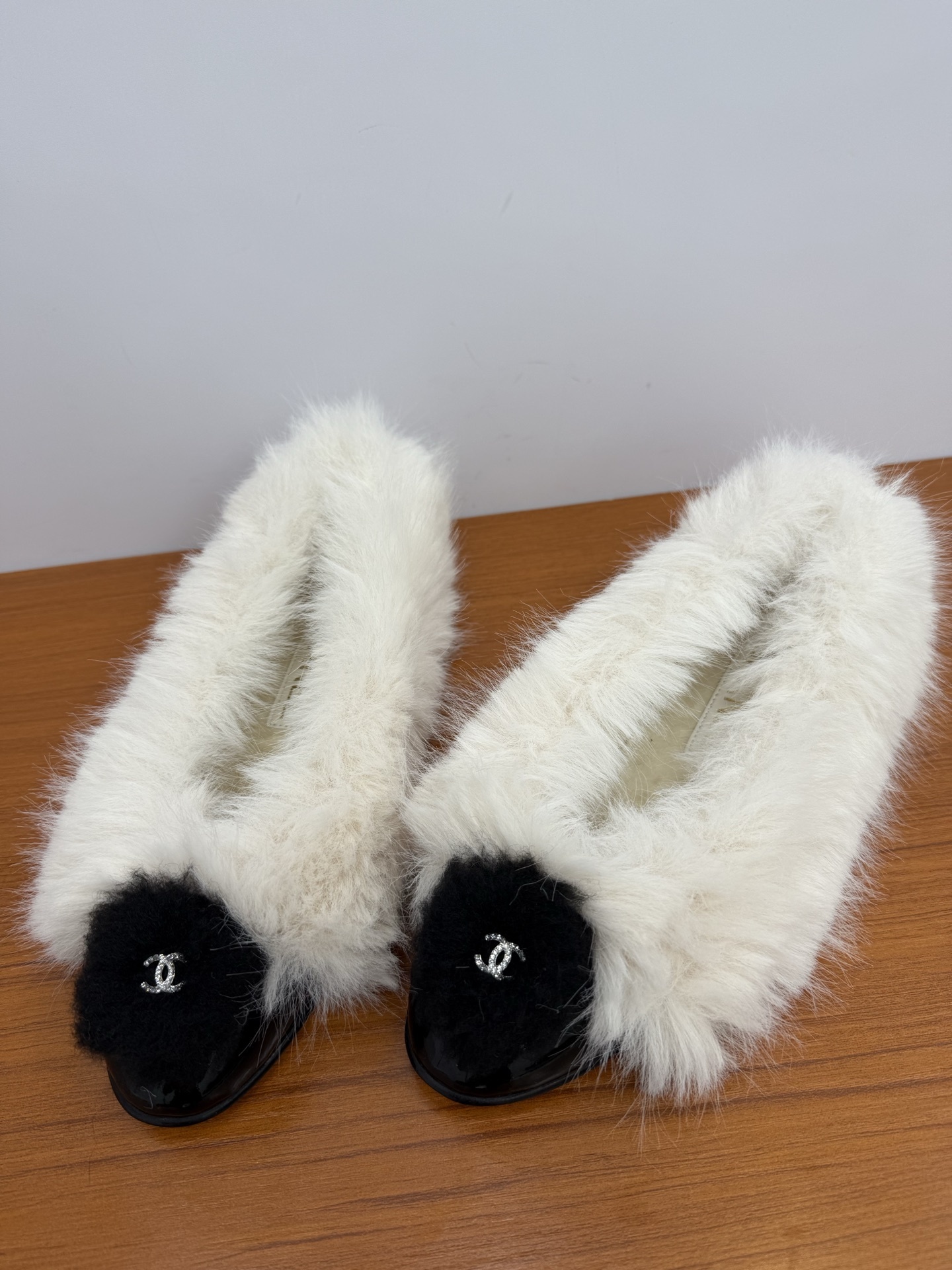 Pair of white faux fur slippers with black fur toe caps and silver crystal CC logos on a wooden surface - Hình 6