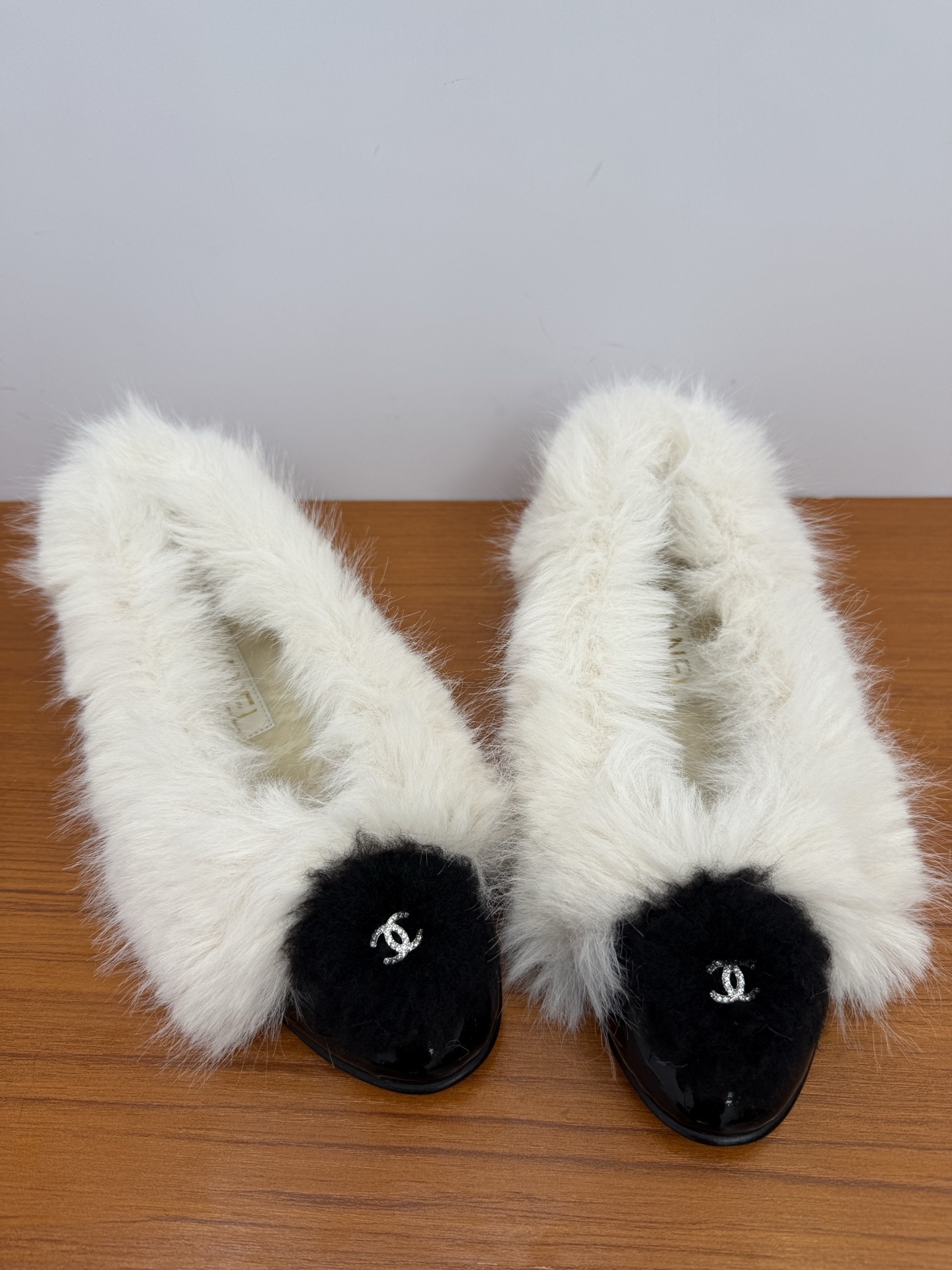 Pair of white faux fur slippers with black fur toe caps and silver crystal CC logos on a wooden surface - Hình 5