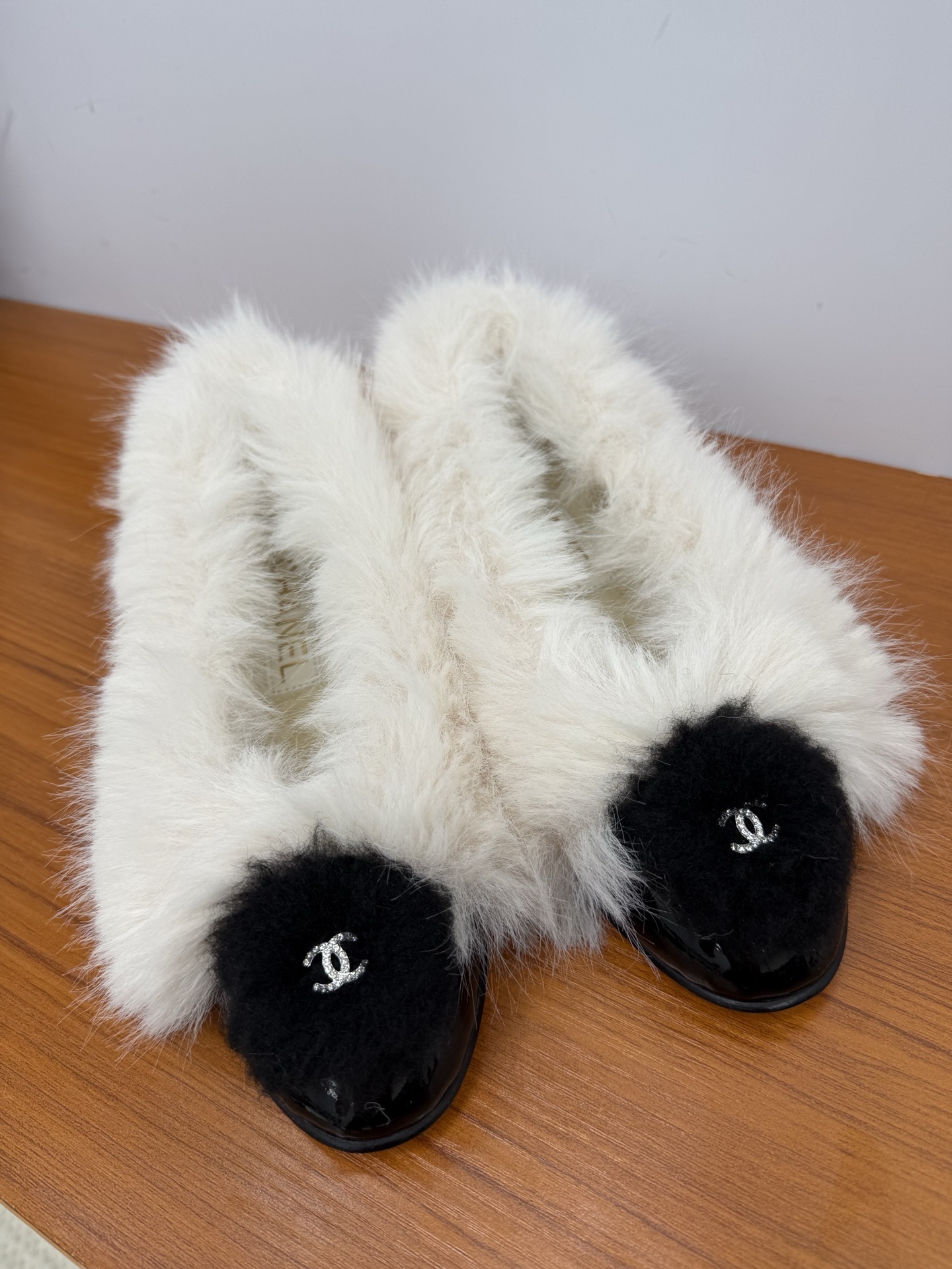 Pair of white faux fur slippers with black fur toe caps and silver crystal CC logos on a wooden surface - Hình 4