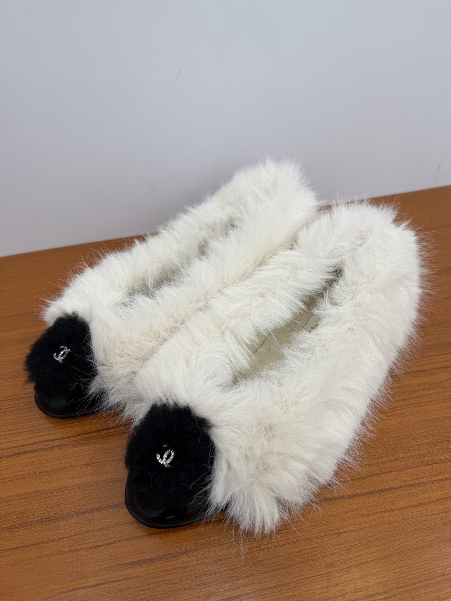 Pair of white faux fur slippers with black fur toe caps and silver crystal CC logos on a wooden surface - Hình 3